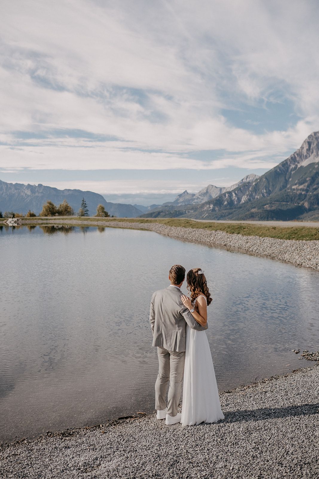 Bride and groom standing by a mountain lake with scenic alpine landscape in background.