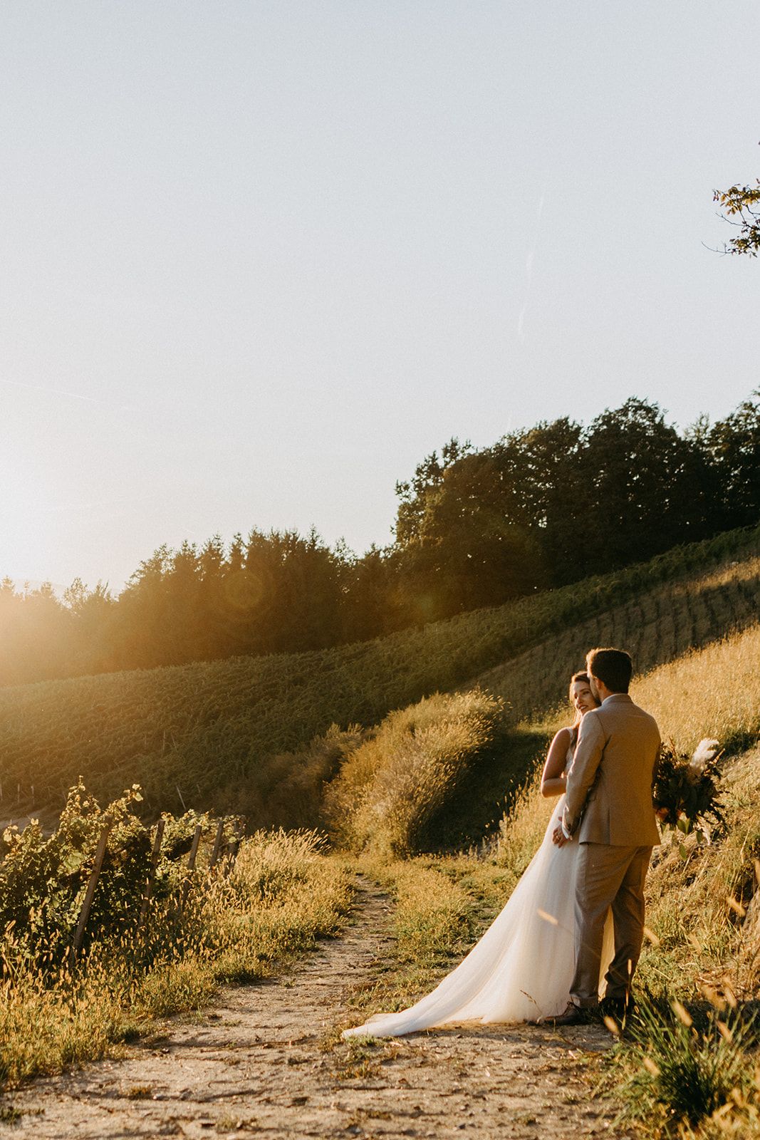 Wedding couple standing together on a rustic path surrounded by golden fields at sunset.