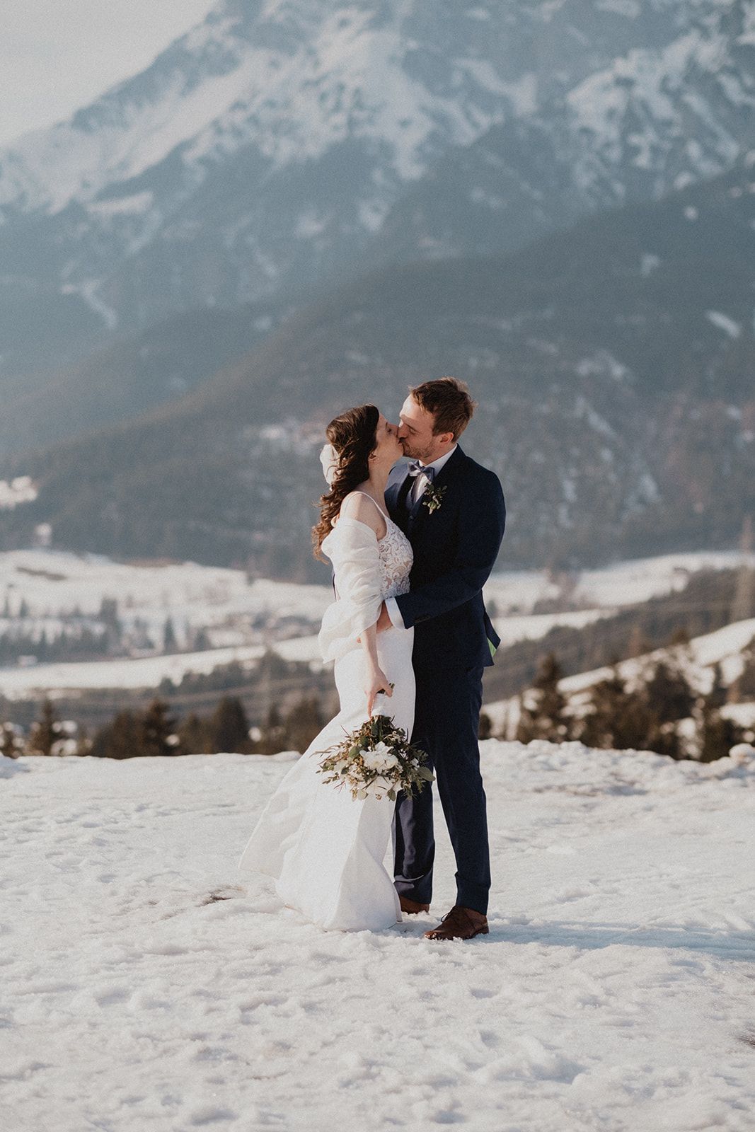 Bride and groom share romantic kiss in snow with majestic mountain landscape behind them at winter wedding. Jufenalm