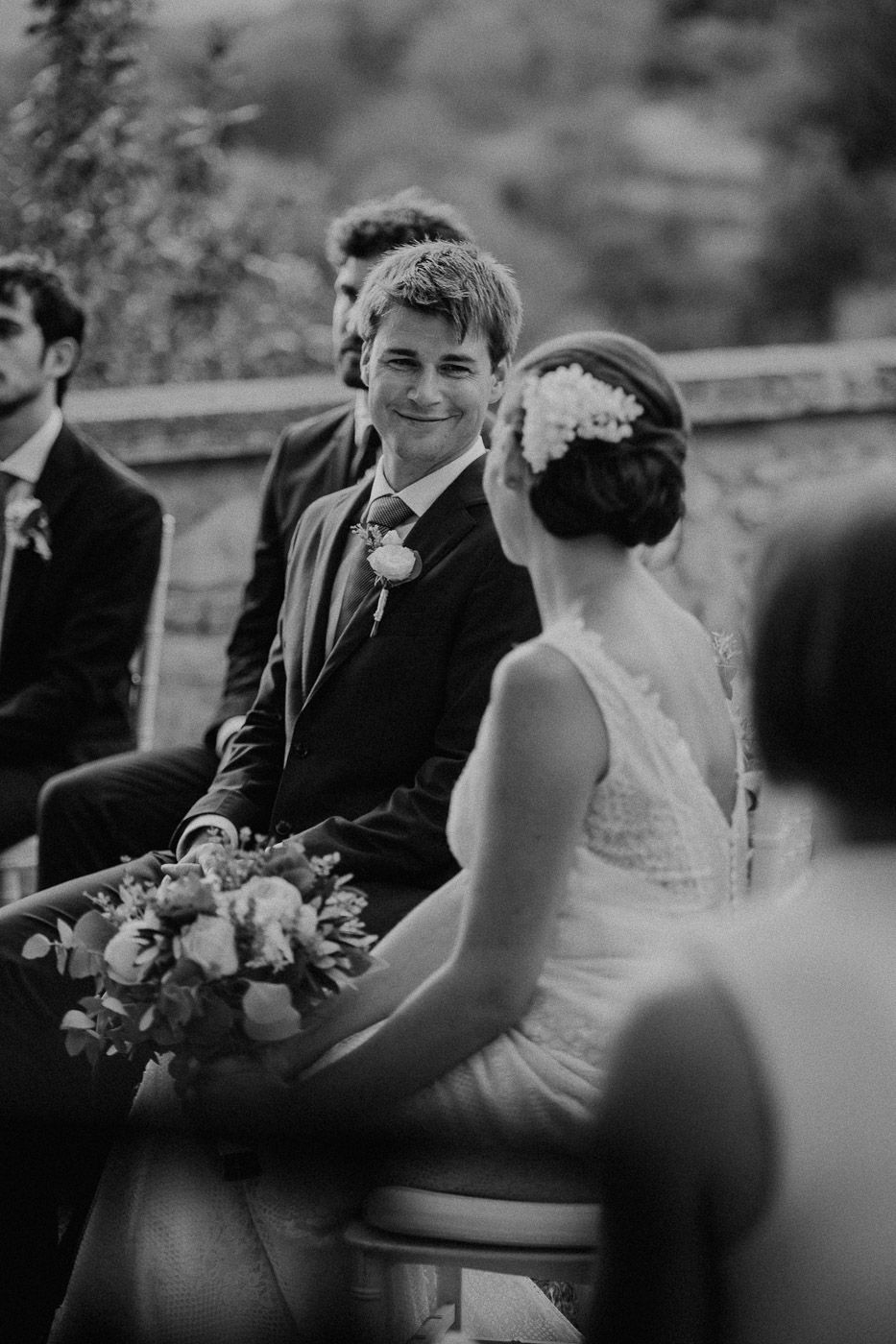 Black and white photo of smiling groom in suit looking at bride during outdoor wedding ceremony. Tuscany Villa Catignano