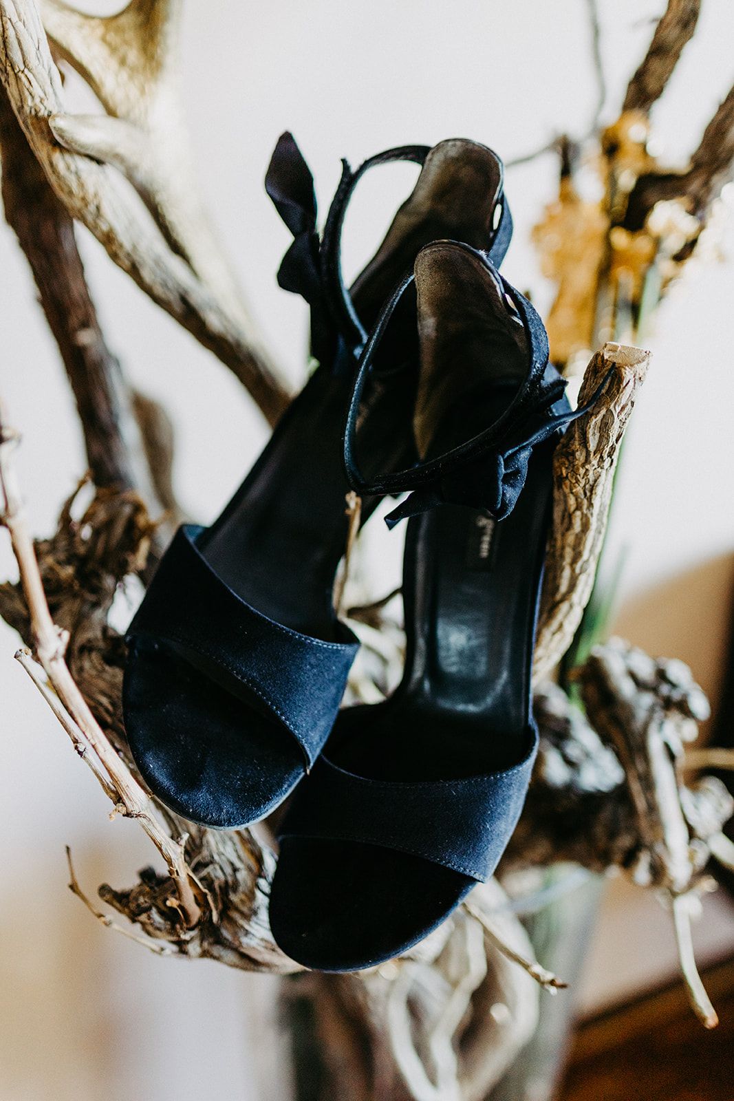 Navy blue suede block heel sandals with ankle strap displayed on rustic driftwood branches.