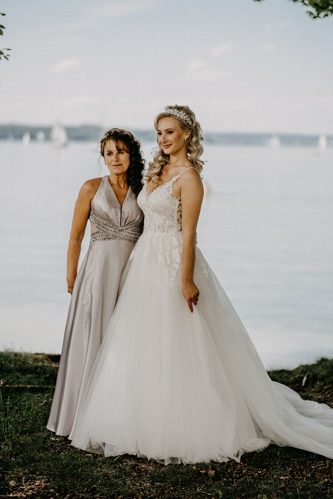 Bride in lace ballgown and mother in silver dress posing together by a lakeside venue.
