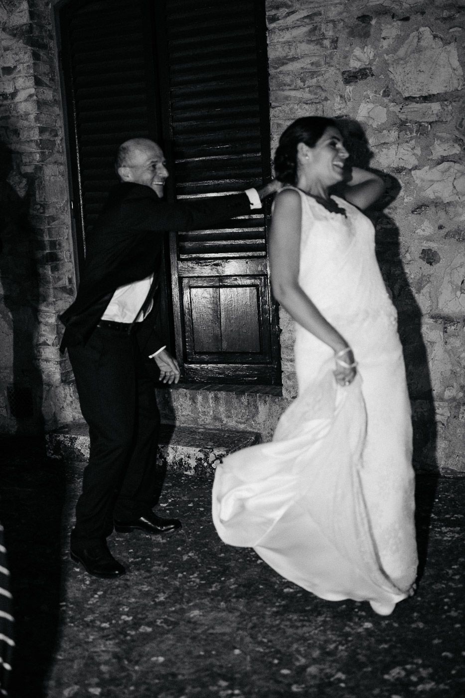 Black and white photo of bride in white gown dancing joyfully with guest at outdoor wedding reception. Tuscany Villa Catignano