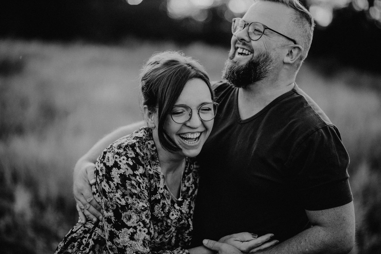 Black and white photo of a laughing couple embracing outdoors in a field during golden hour.