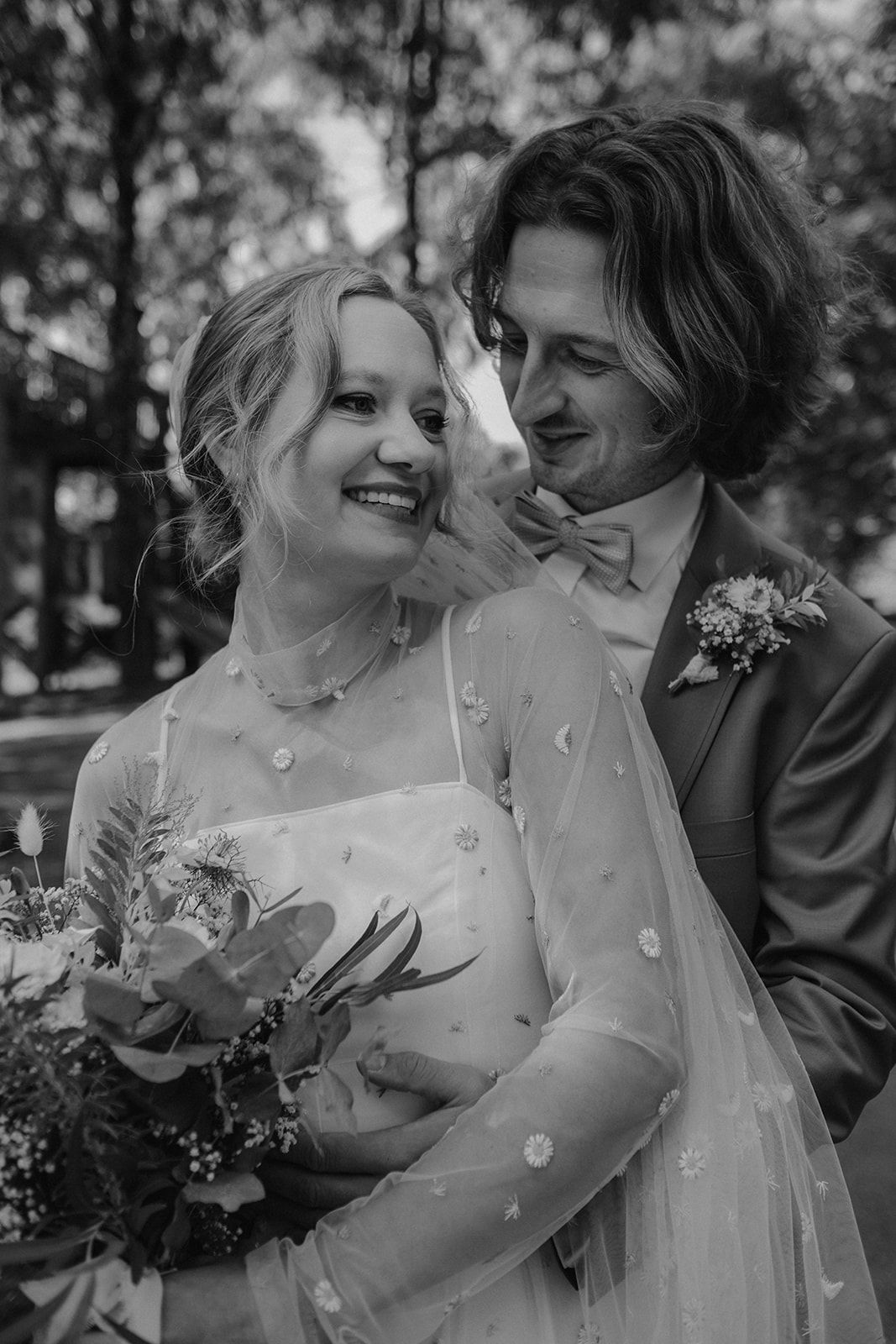 Black and white photo of smiling bride in floral veil held by groom in bow tie and boutonniere. Gut Dietersberg
