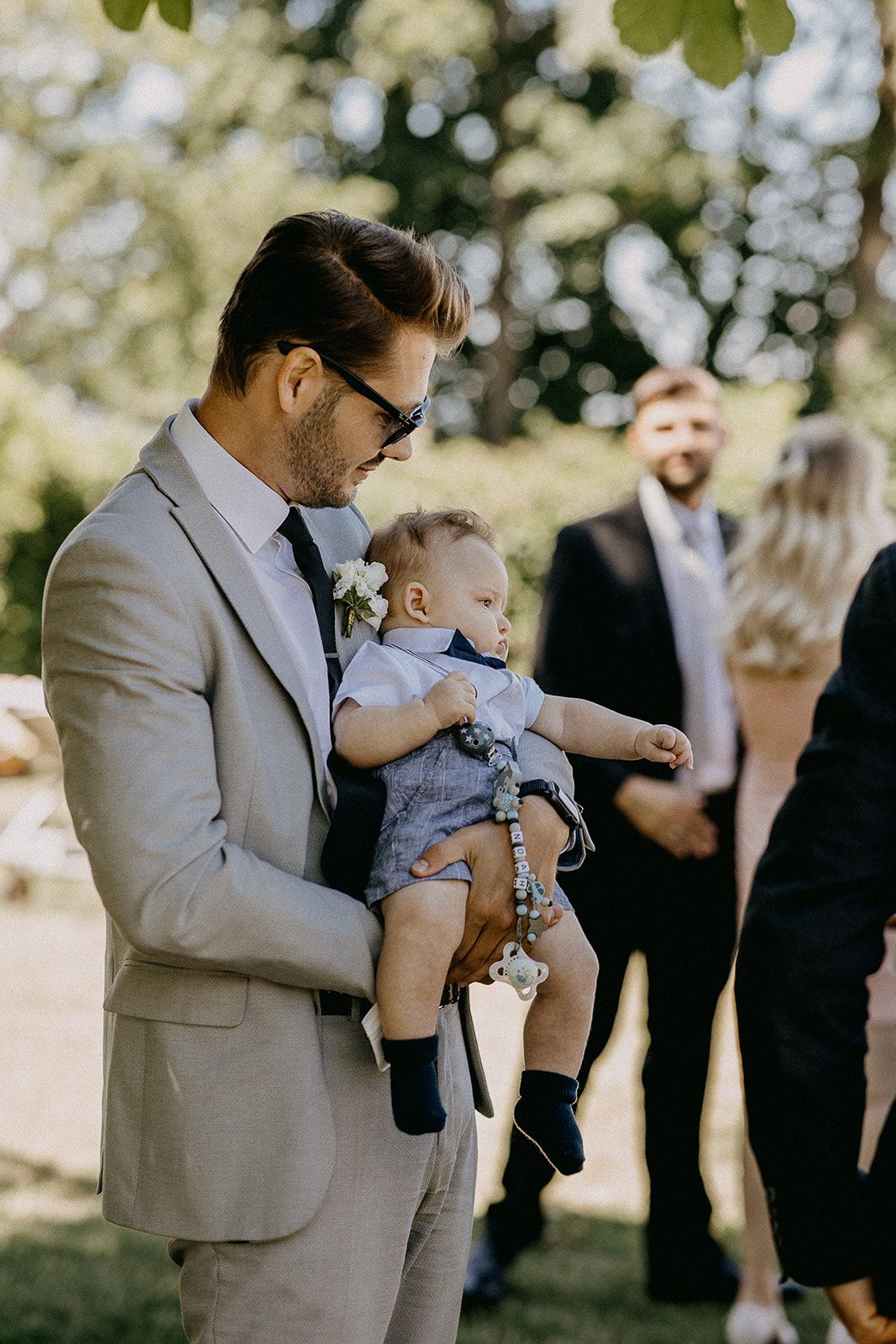 A well-dressed man in a beige suit holds a baby during an outdoor wedding ceremony in sunlight.