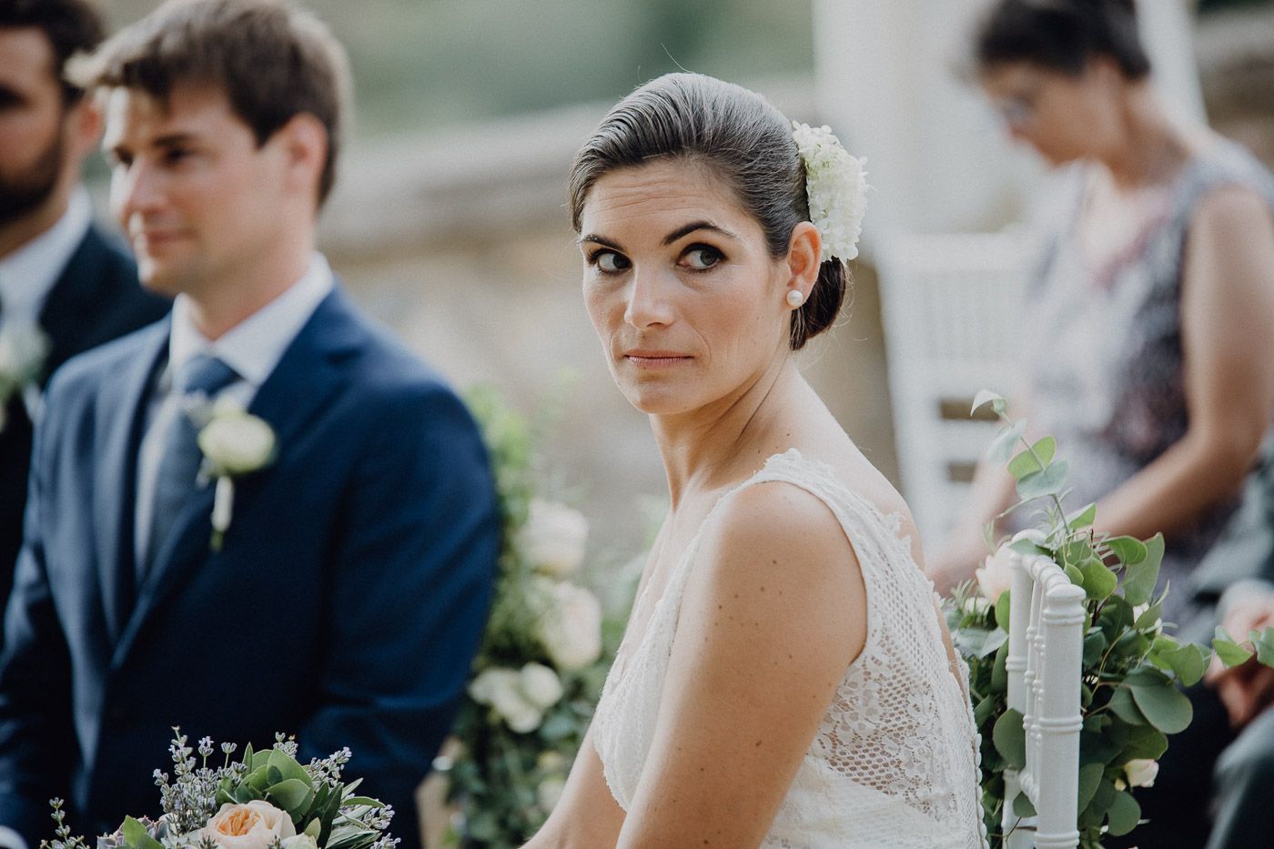 Bride in white lace dress looks back over shoulder during outdoor wedding ceremony with floral decorations. Tuscany Villa Catignano