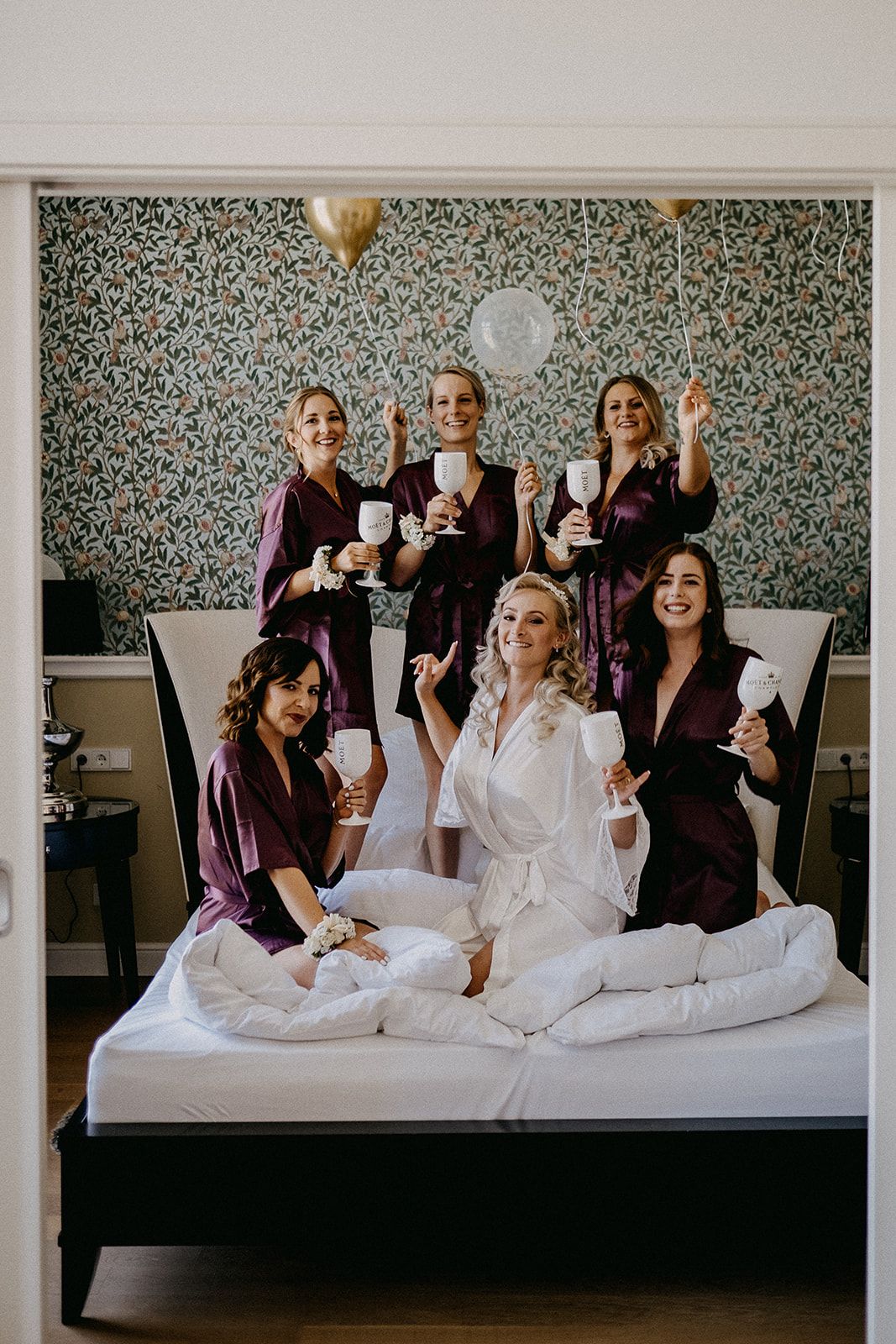 Bride in white robe with five bridesmaids in burgundy robes celebrating on a bed with champagne glasses.