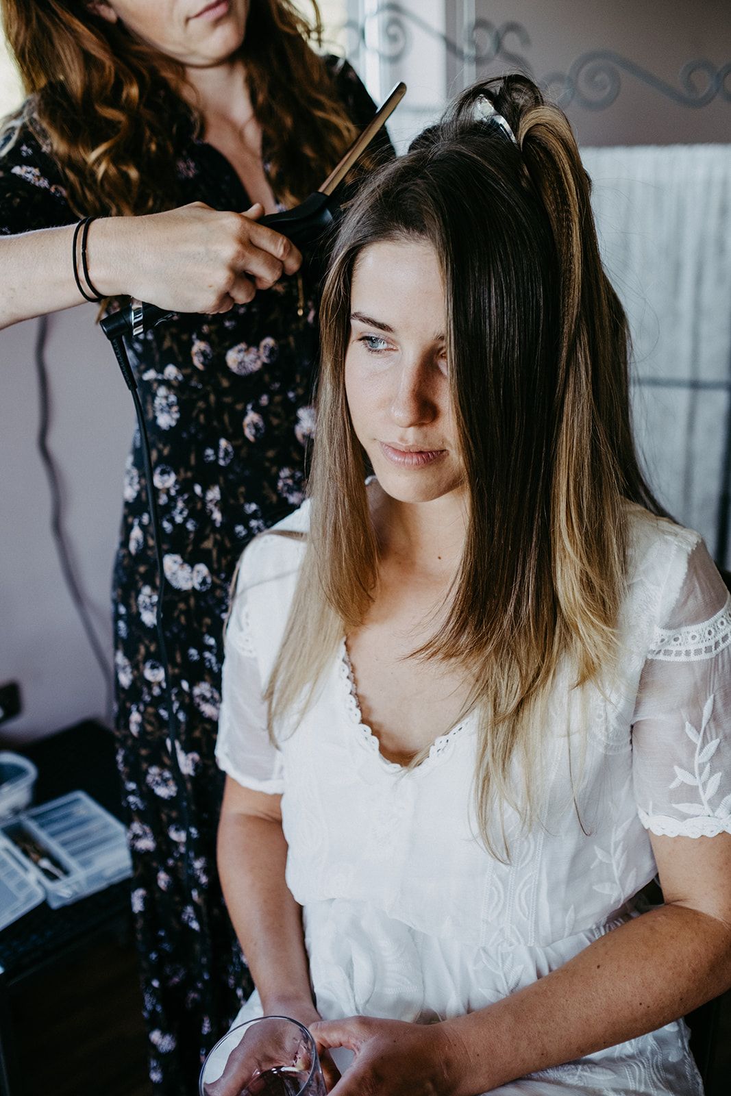 Hair stylist in floral dress styling brunette brides hair during wedding preparations.