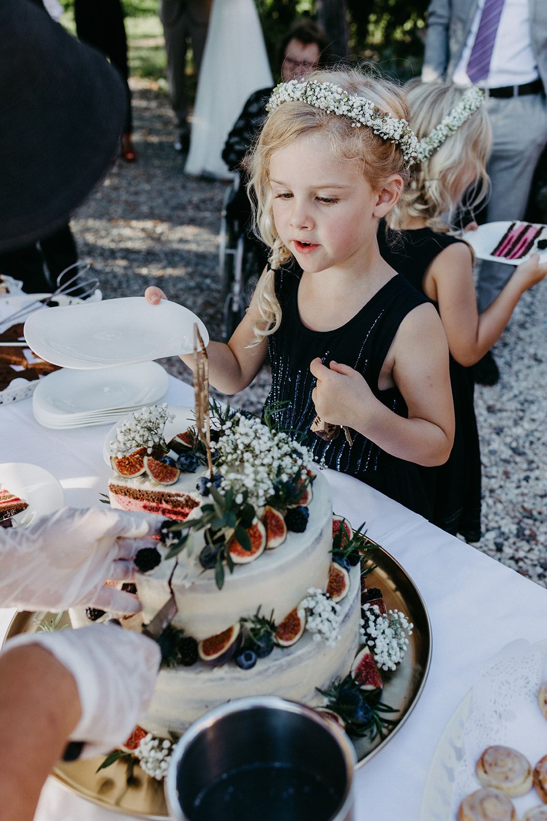 Young flower girl in black dress with floral crown watching wedding cake being cut at outdoor reception.