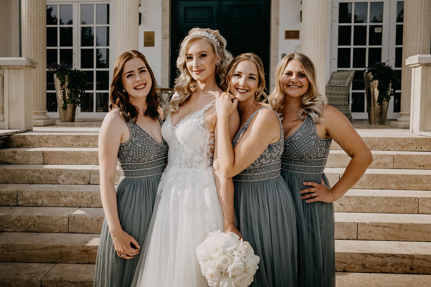 Bride in lace wedding gown poses with three bridesmaids in gray dresses holding white bouquet.