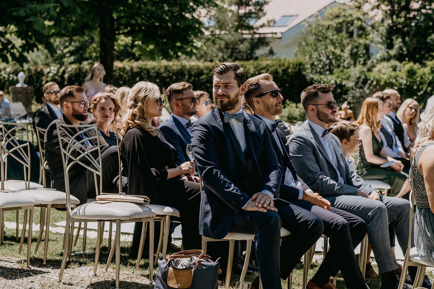 Smartly dressed wedding guests in navy suits smile and enjoy outdoor garden wedding ceremony.