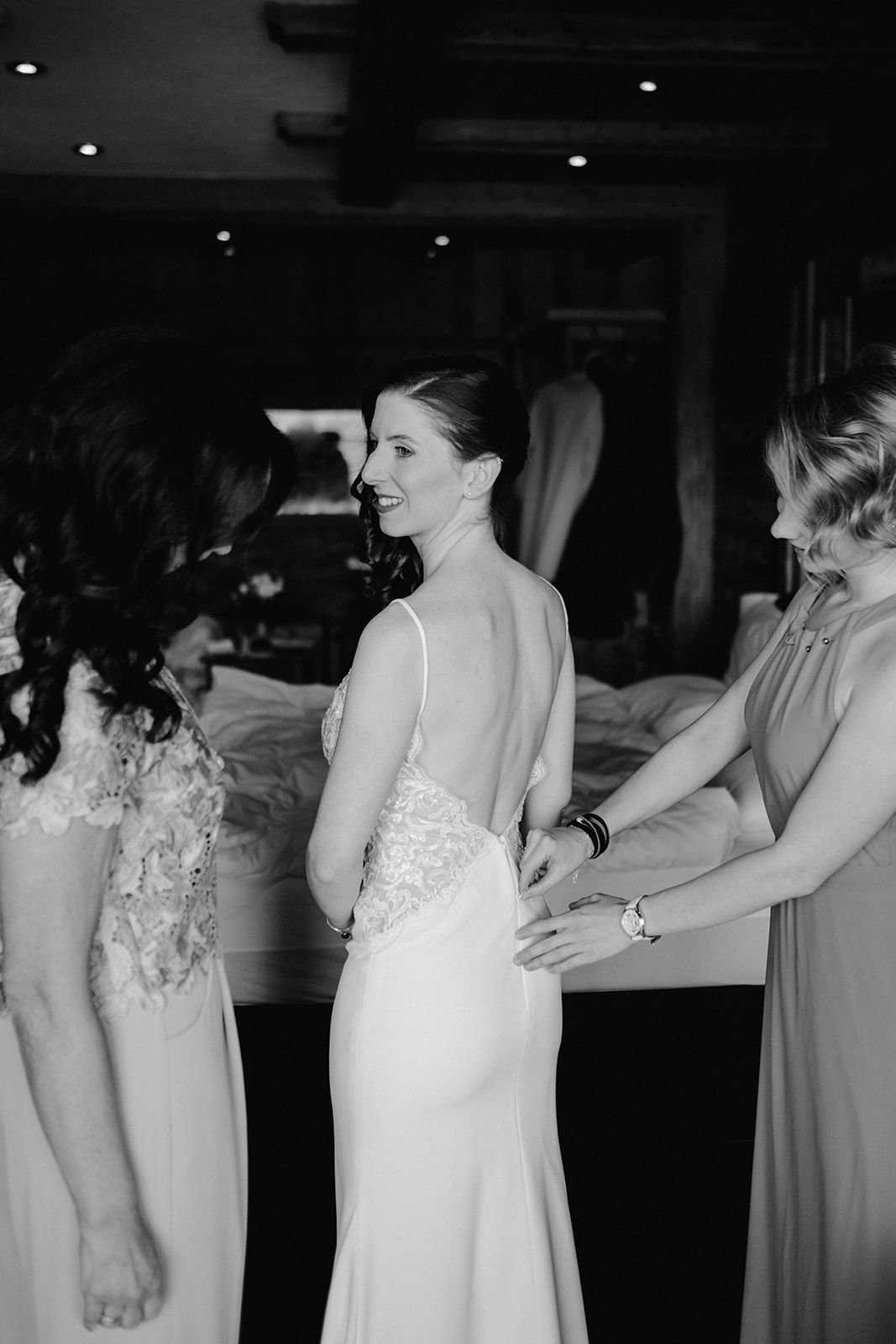 Bride smiling over shoulder in backless lace wedding gown while bridesmaids help her dress.