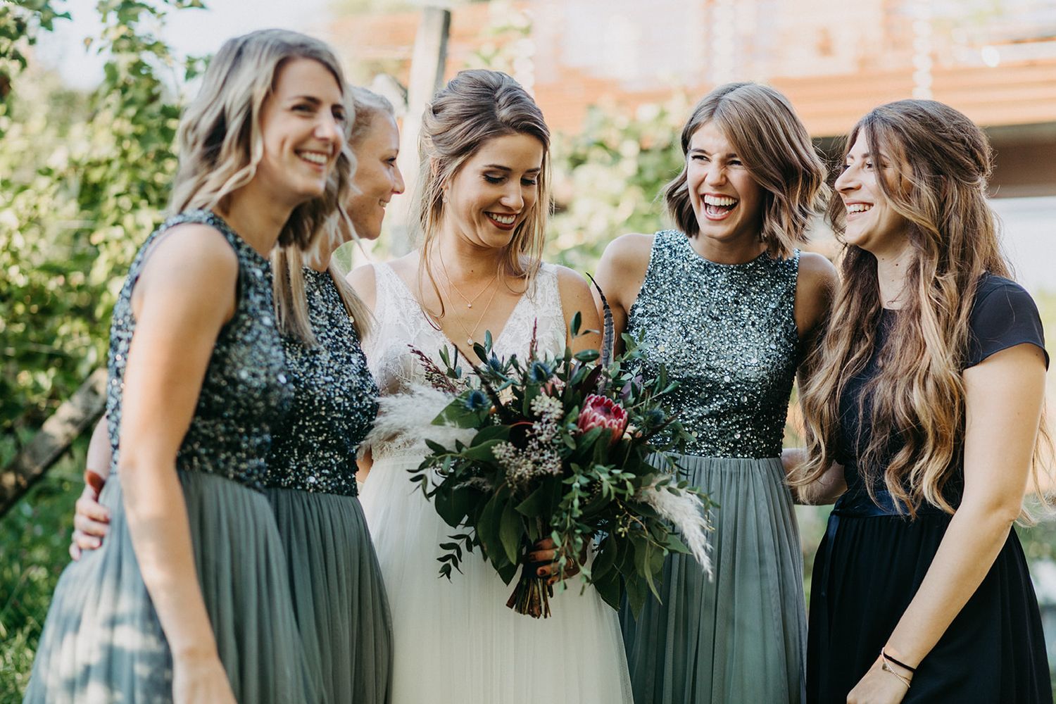 Bride surrounded by bridesmaids in matching silver sequin tops and sage green skirts holding a bouquet.
