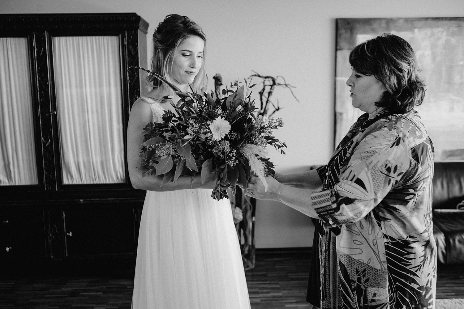 Bride receiving bouquet of flowers from woman in black and white wedding preparation moment.