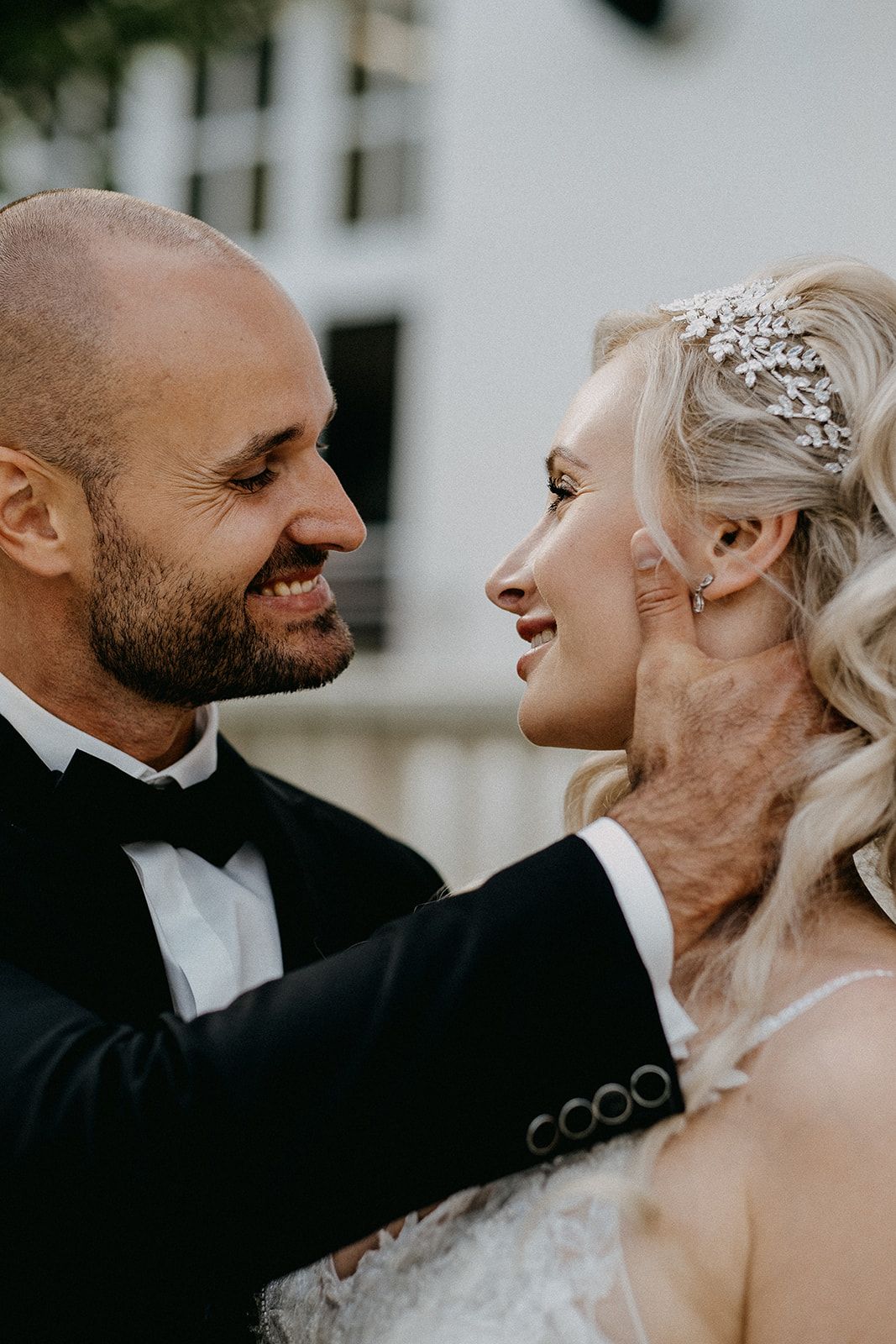 Smiling bride and groom share an intimate moment, she wears a crystal hair piece and lace gown.