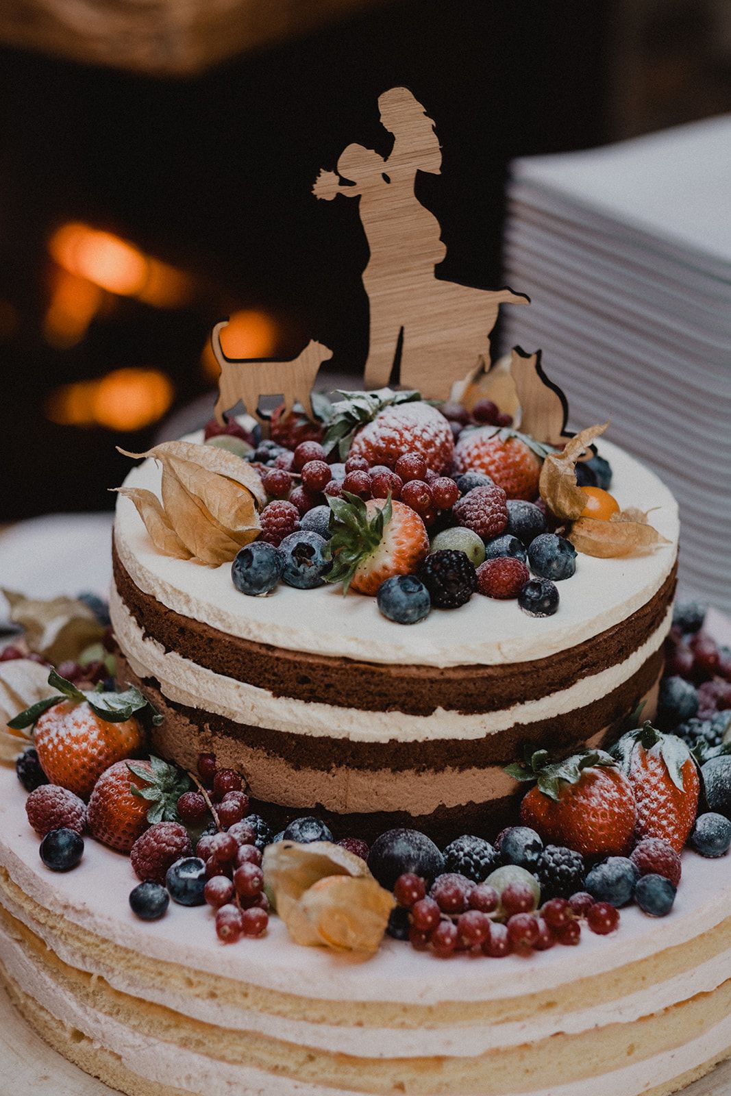 Two-tier naked wedding cake topped with fresh berries, strawberries, blueberries and wooden bride and groom silhouette topper. Jufenalm
