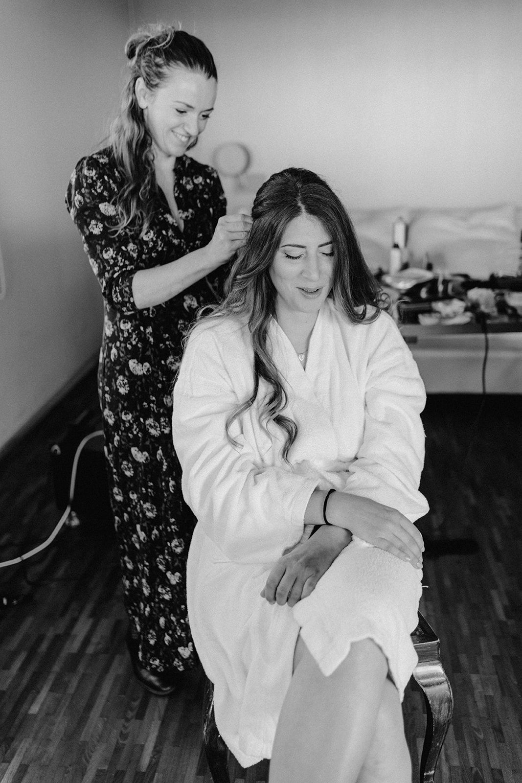 Black and white photo of bride in white robe having her hair styled before wedding ceremony.