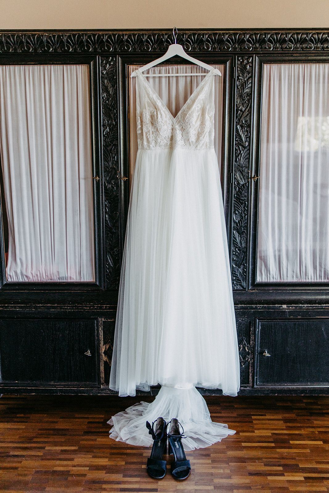 Elegant white lace wedding gown hanging on ornate dark wardrobe with black heels below.
