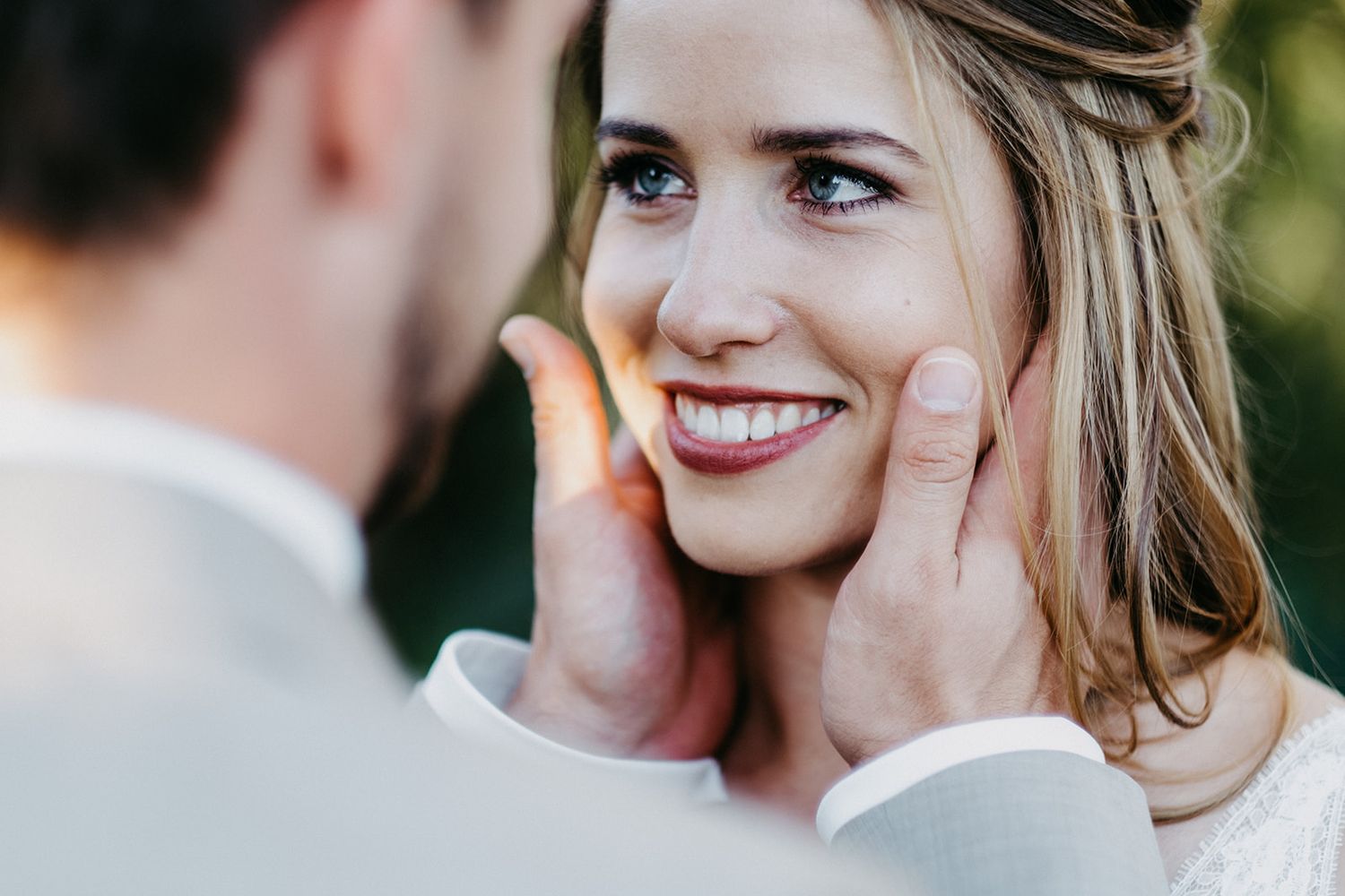 Groom gently cupping smiling bride's face as she gazes up at him with blue eyes in a soft outdoor portrait.