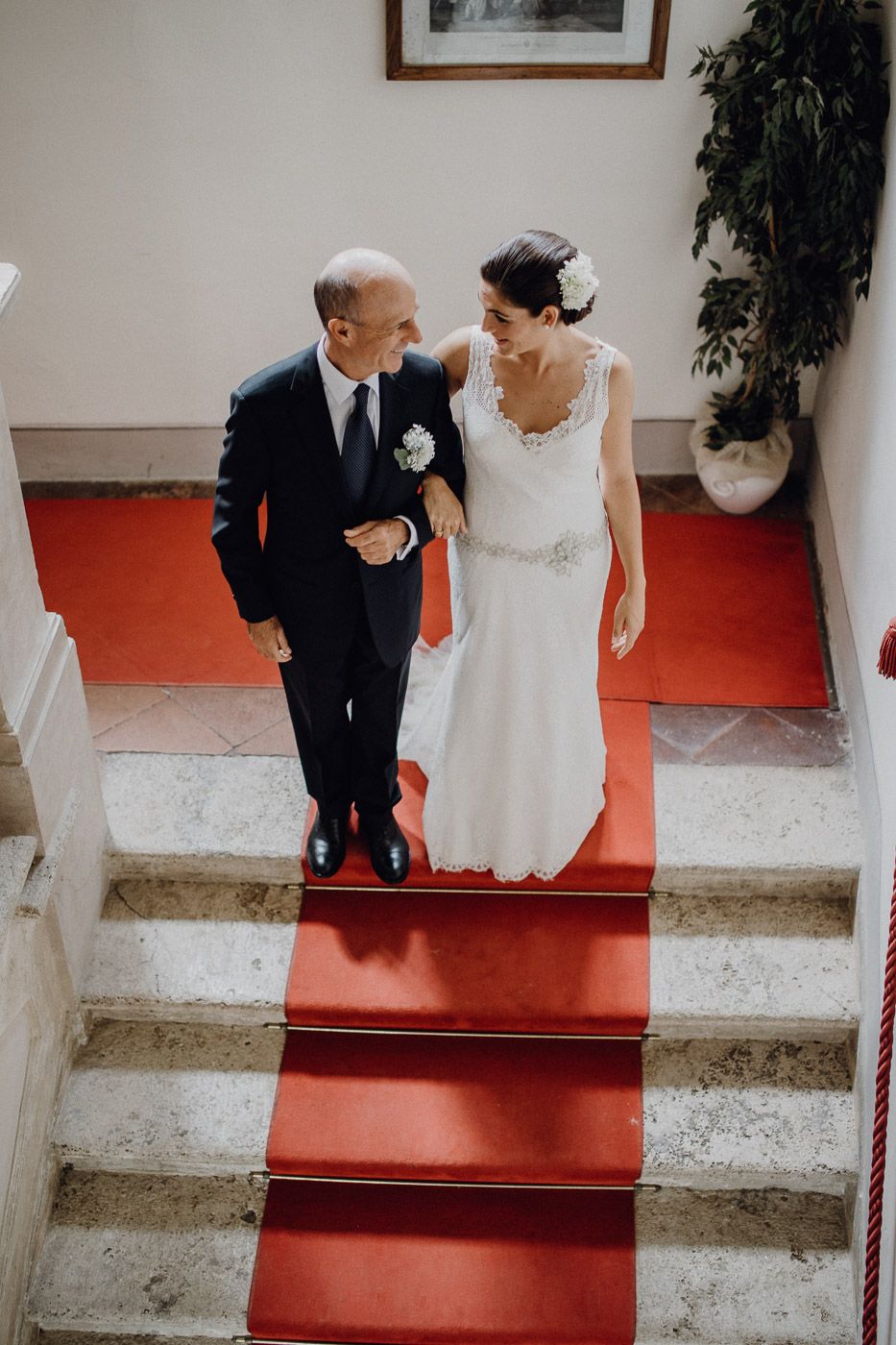 Bride and escort share a joyful moment walking down red-carpeted marble staircase at wedding venue. Tuscany Villa Catignano