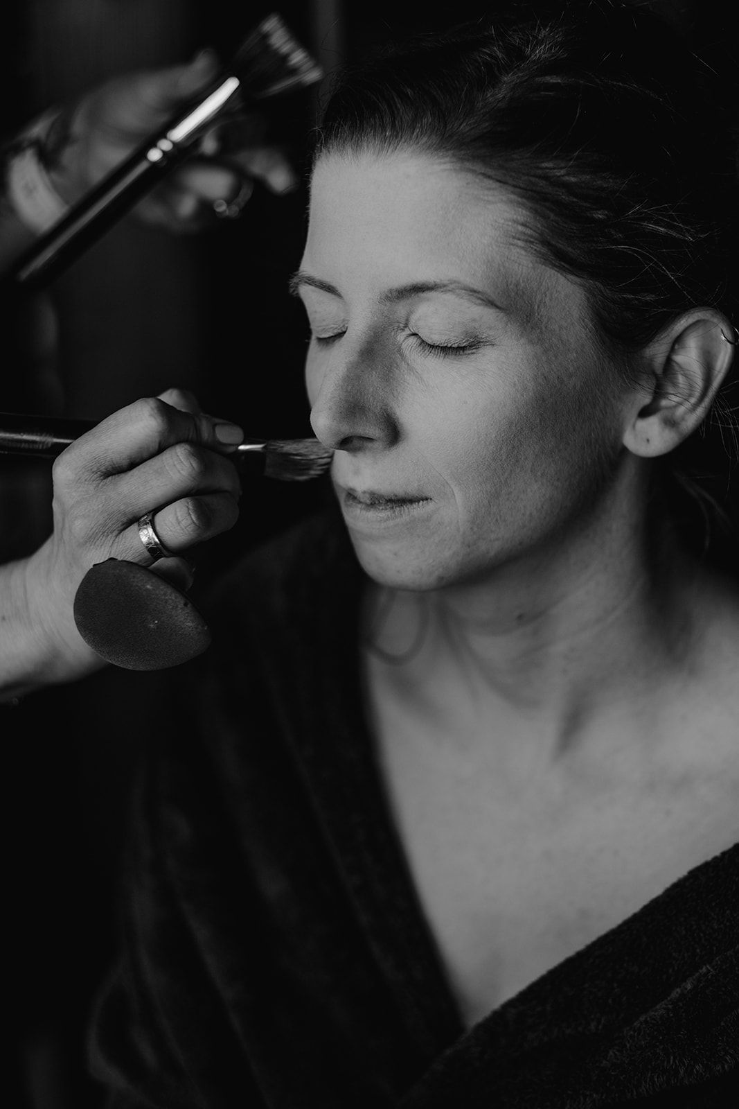 Black and white close-up of a makeup artist applying foundation to a womans face with a brush.