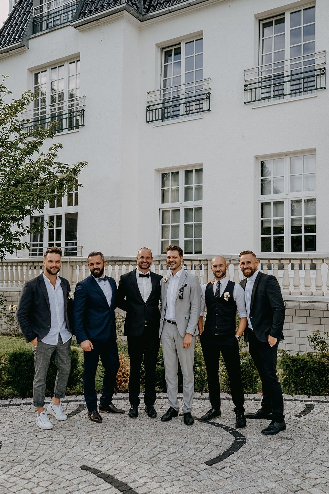 Groom in tuxedo poses with six groomsmen in suits outside elegant white wedding venue.