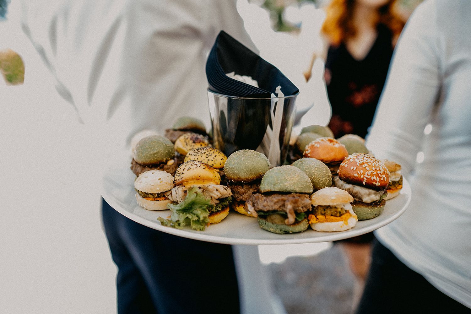 A waiter holds a large platter of assorted mini sliders and small burgers at a catered wedding reception event.