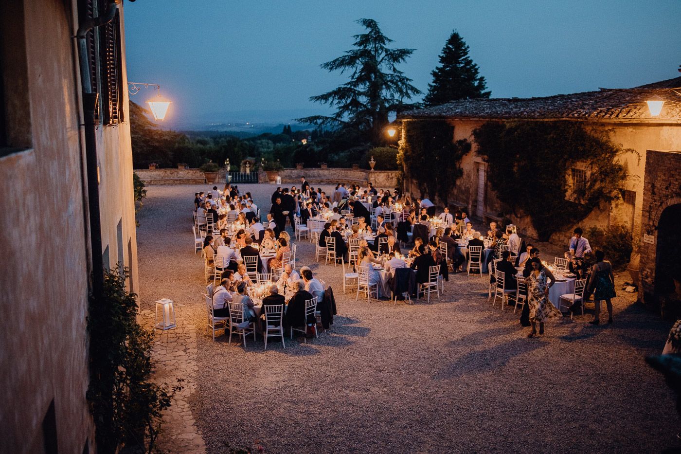 Aerial view of wedding guests dining at candlelit tables in Italian villa courtyard at dusk. Tuscany Villa Catignano
