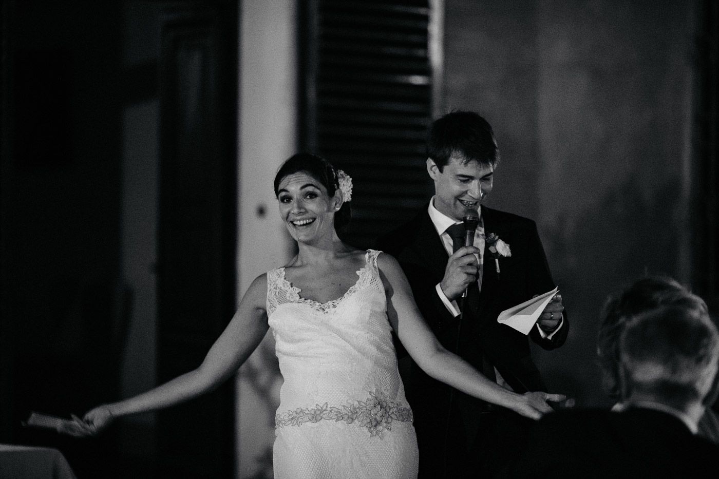 Smiling bride in lace gown and groom with microphone during wedding speech in black and white. Tuscany Villa Catignano