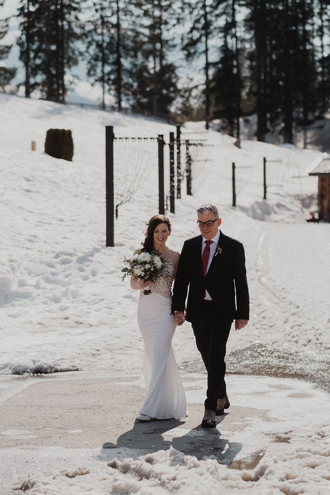Bride and groom walking hand in hand through snowy winter landscape with wedding bouquet.