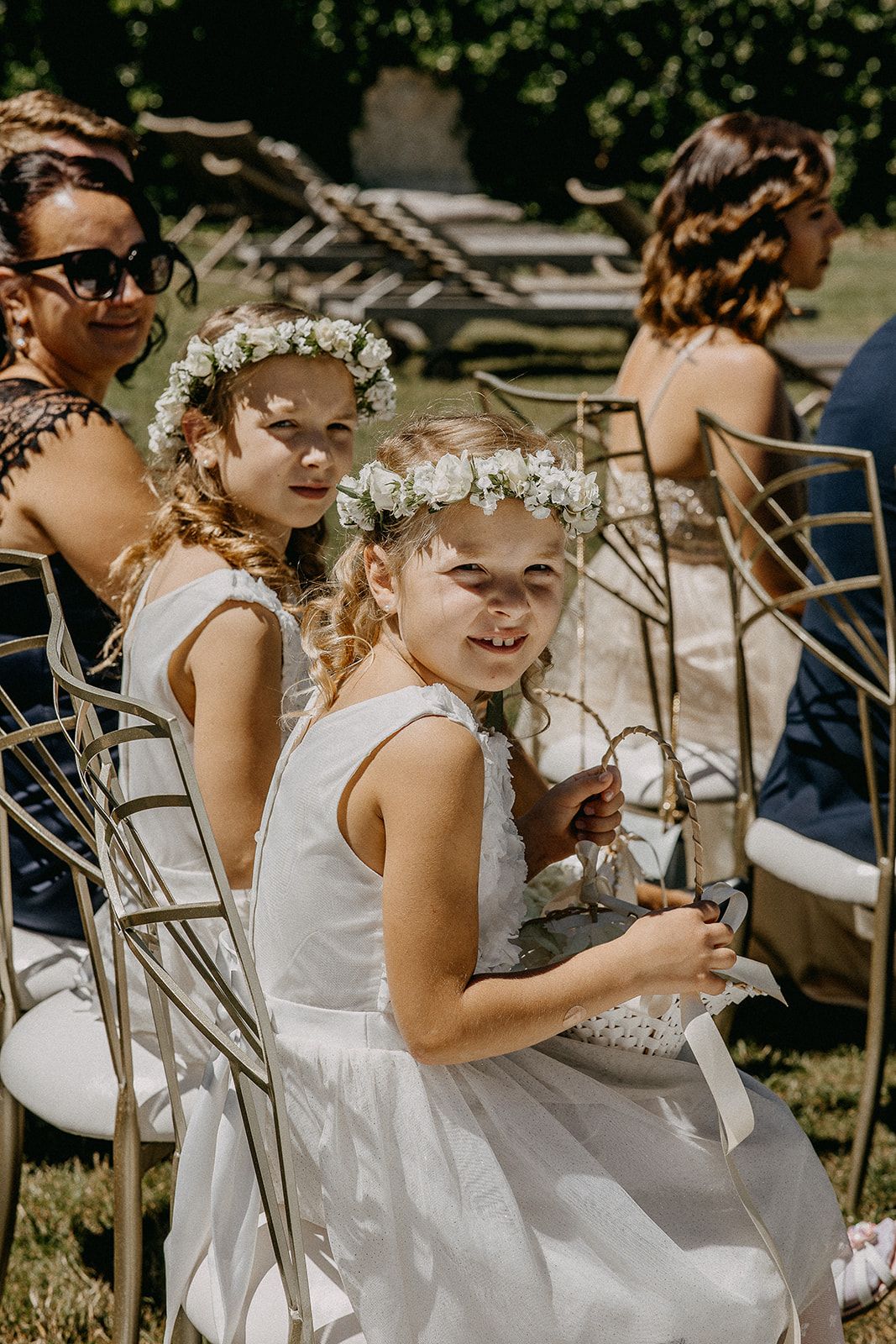 Two young flower girls in white dresses and floral crowns sit smiling during an outdoor wedding ceremony.