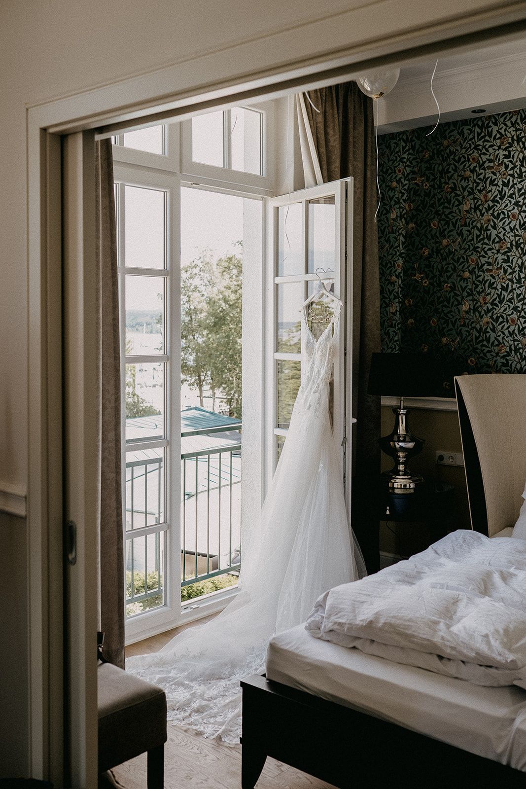 Flowing white wedding dress with long veil hanging by open French doors with water view.