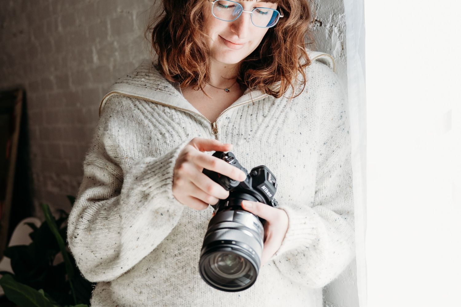 Person in white sweater adjusting camera lens near bright window.