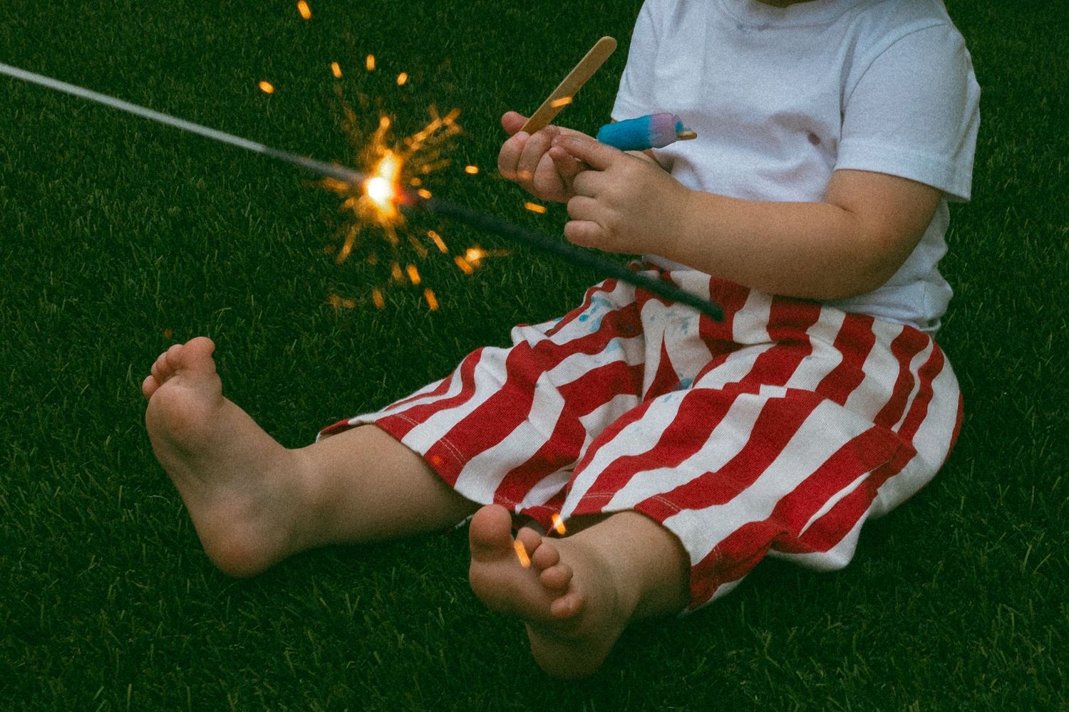 young child sitting in the grass holding a popsicle with a sparkler nearby