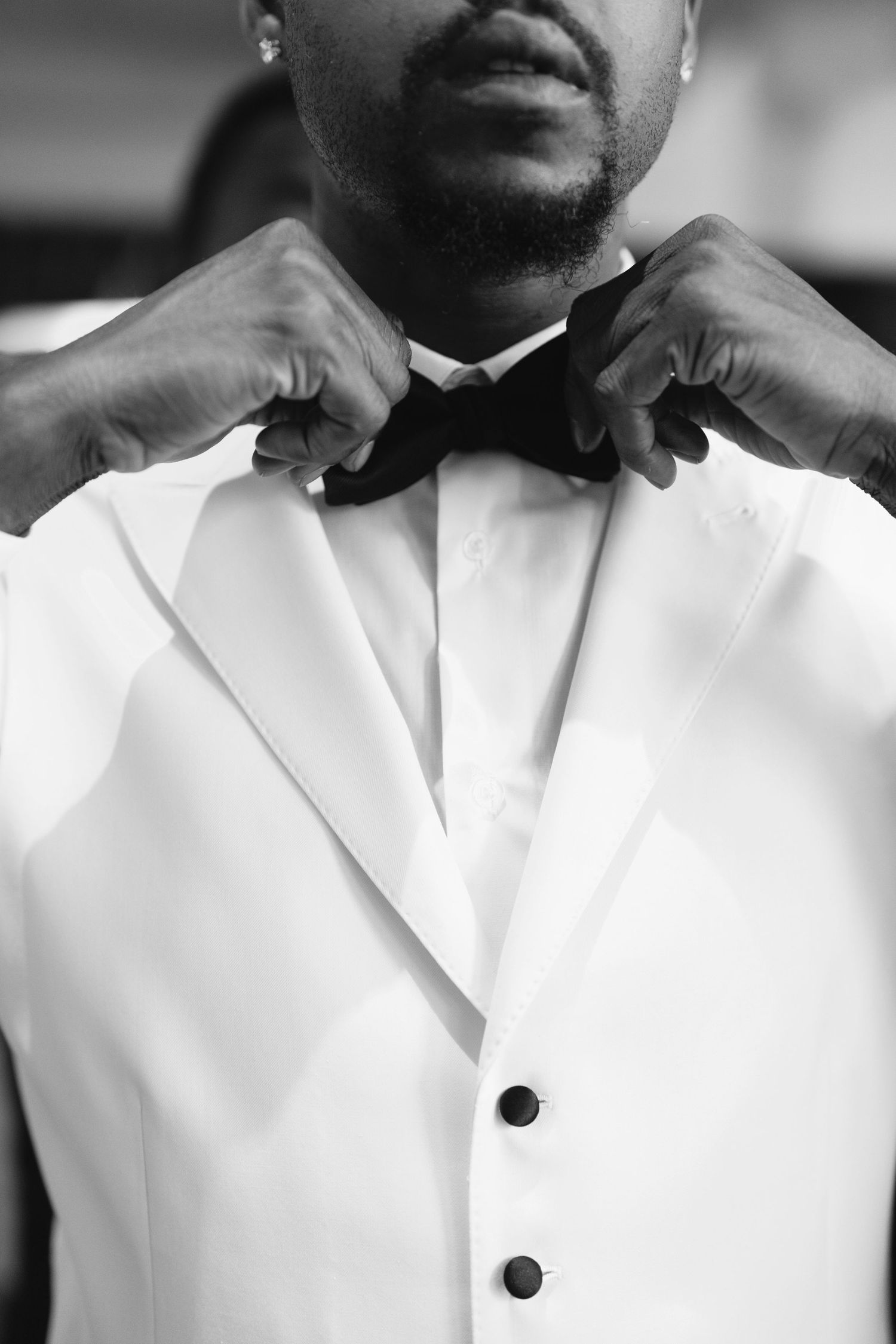 Atlanta wedding photography of a groom in a white tuxedo adjusting his black bow tie in a black-and-white portrait.