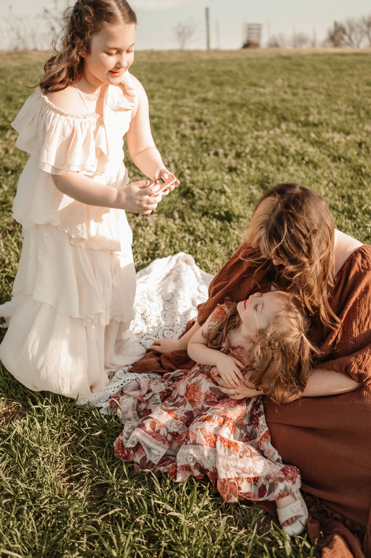 A mother in a brown dress tenderly holds a toddler on grass while an older girl in white offers a flower.