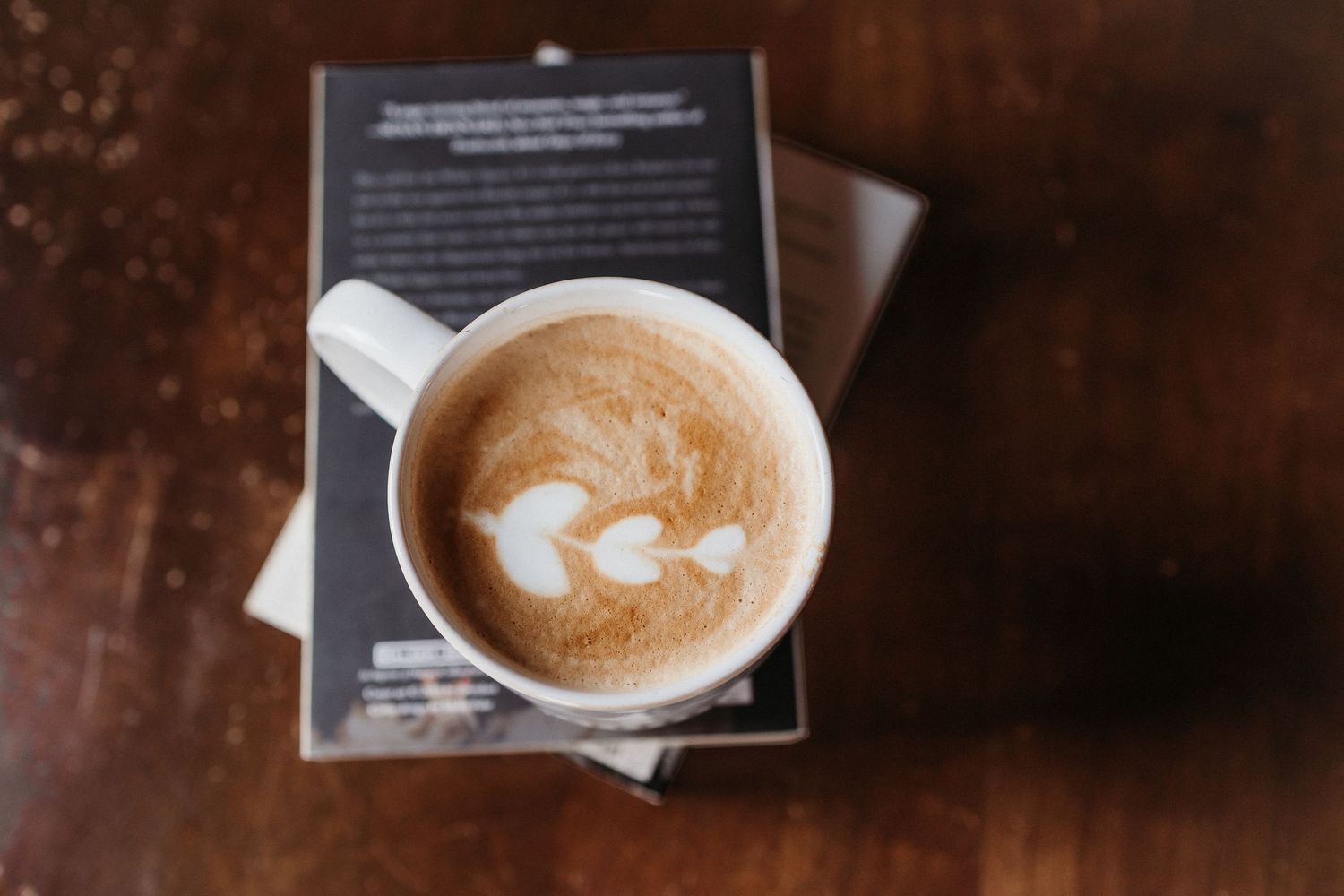 A latte with heart-shaped latte art in a white mug resting on a book on a wooden table.