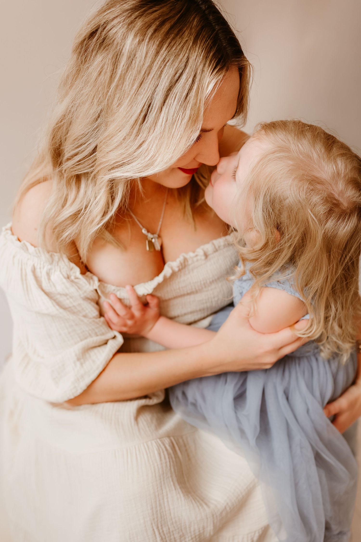 A blonde mother tenderly touches noses with her toddler daughter in matching white and blue dresses.