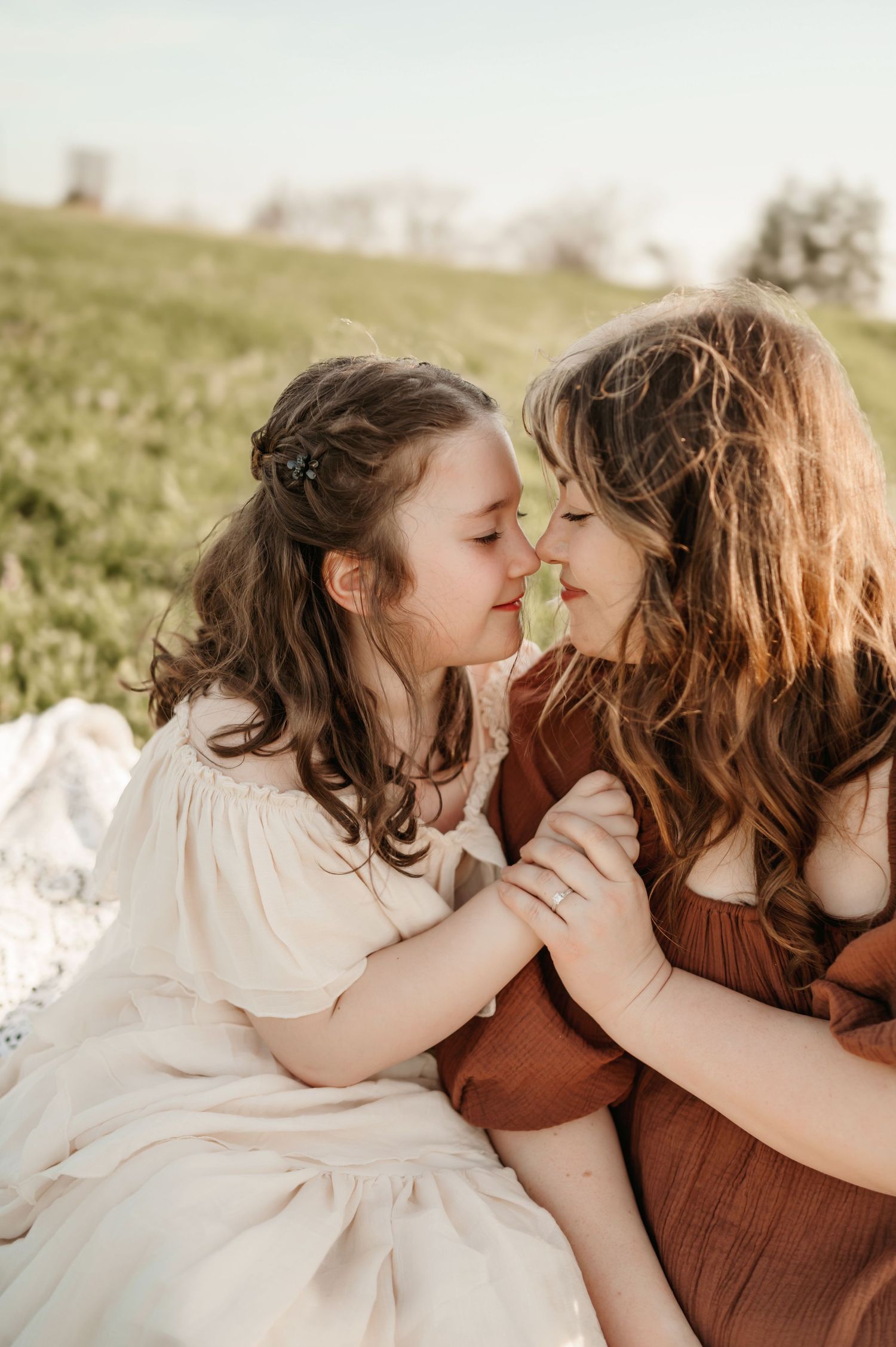 A mother and young daughter share a tender nose-to-nose moment while sitting on a blanket in a green field.