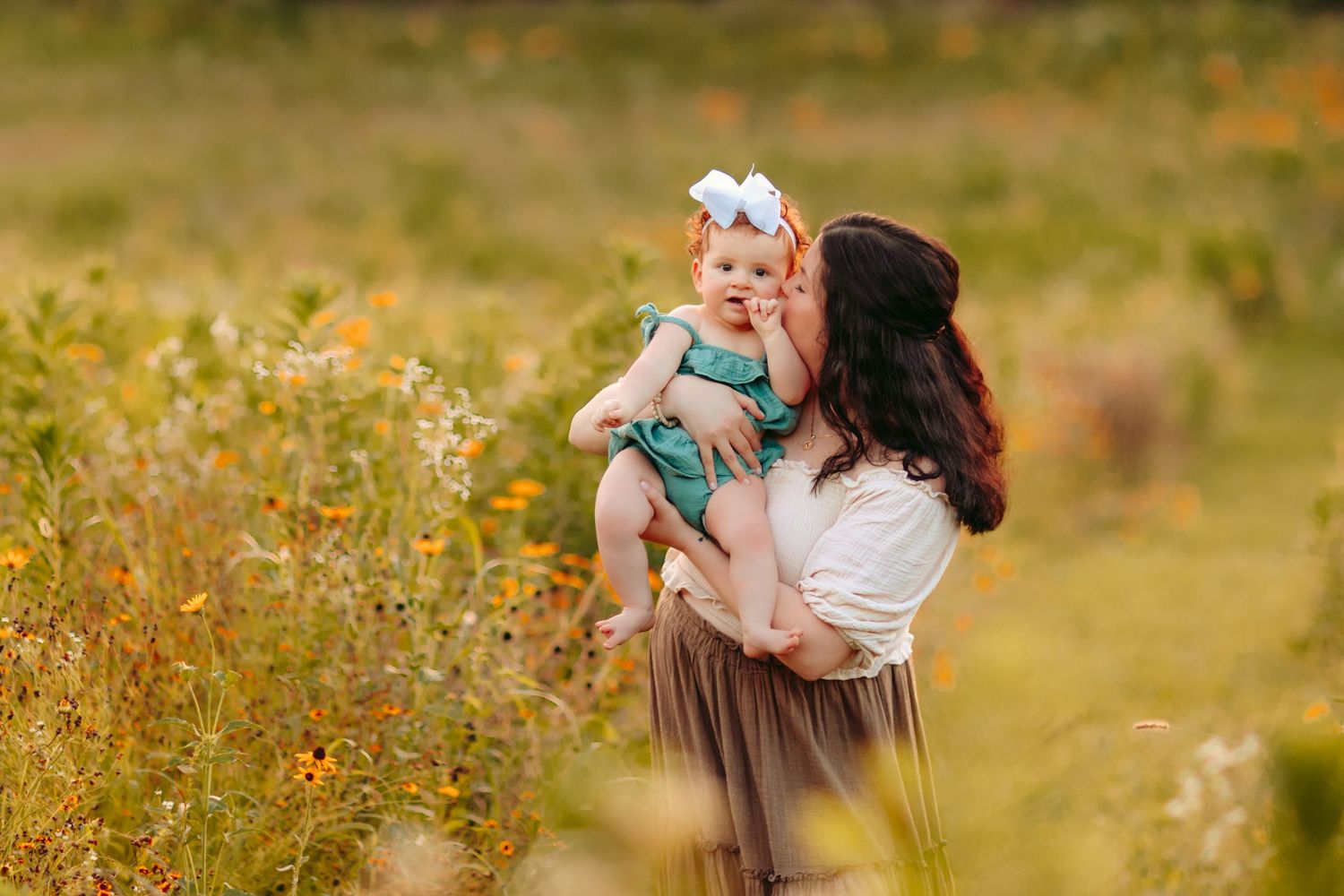 A mother holds her baby girl with a white bow in a sunlit wildflower field, sharing a tender kiss outdoors.