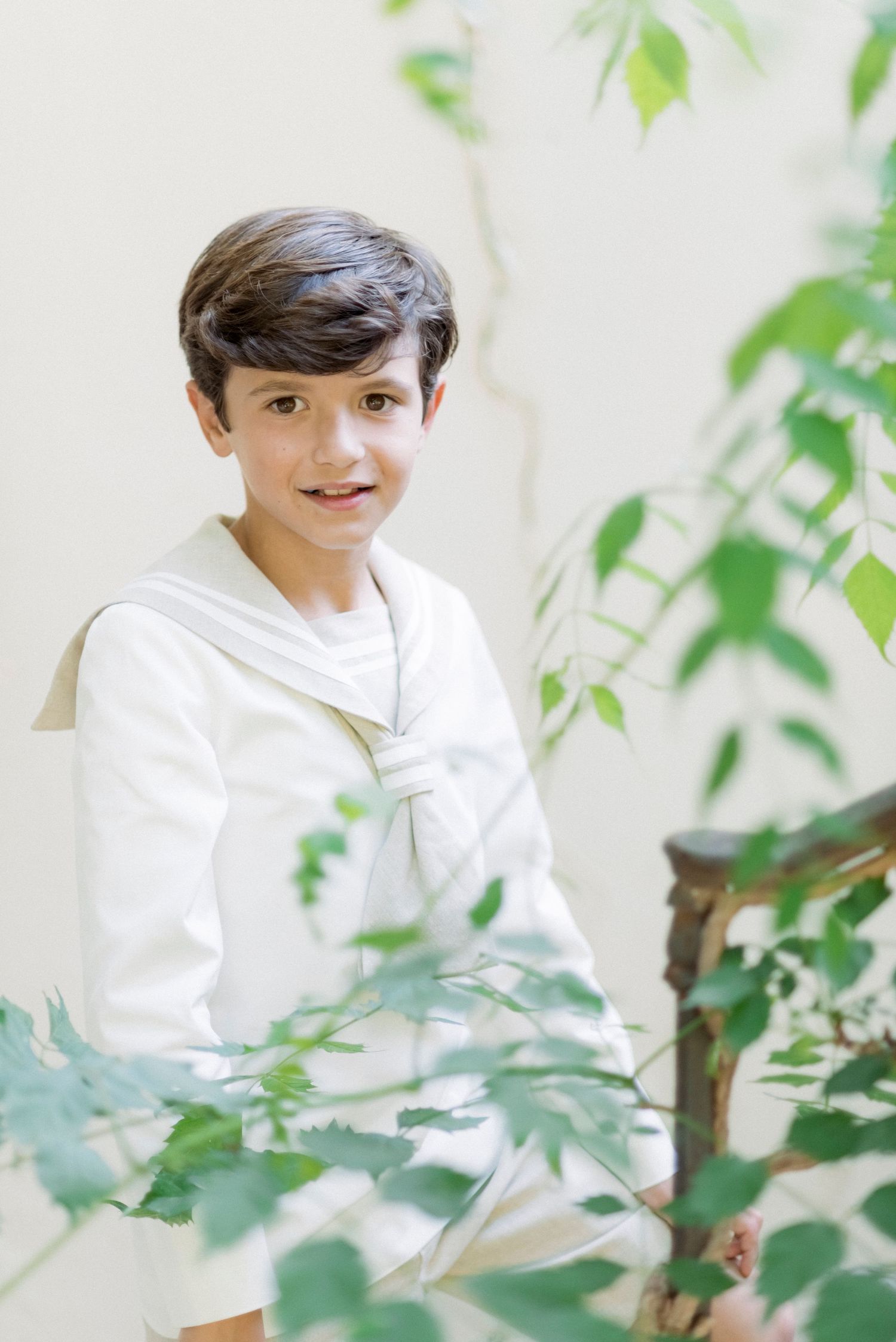 Smiling boy in white sailor communion outfit seated among green leafy plants.