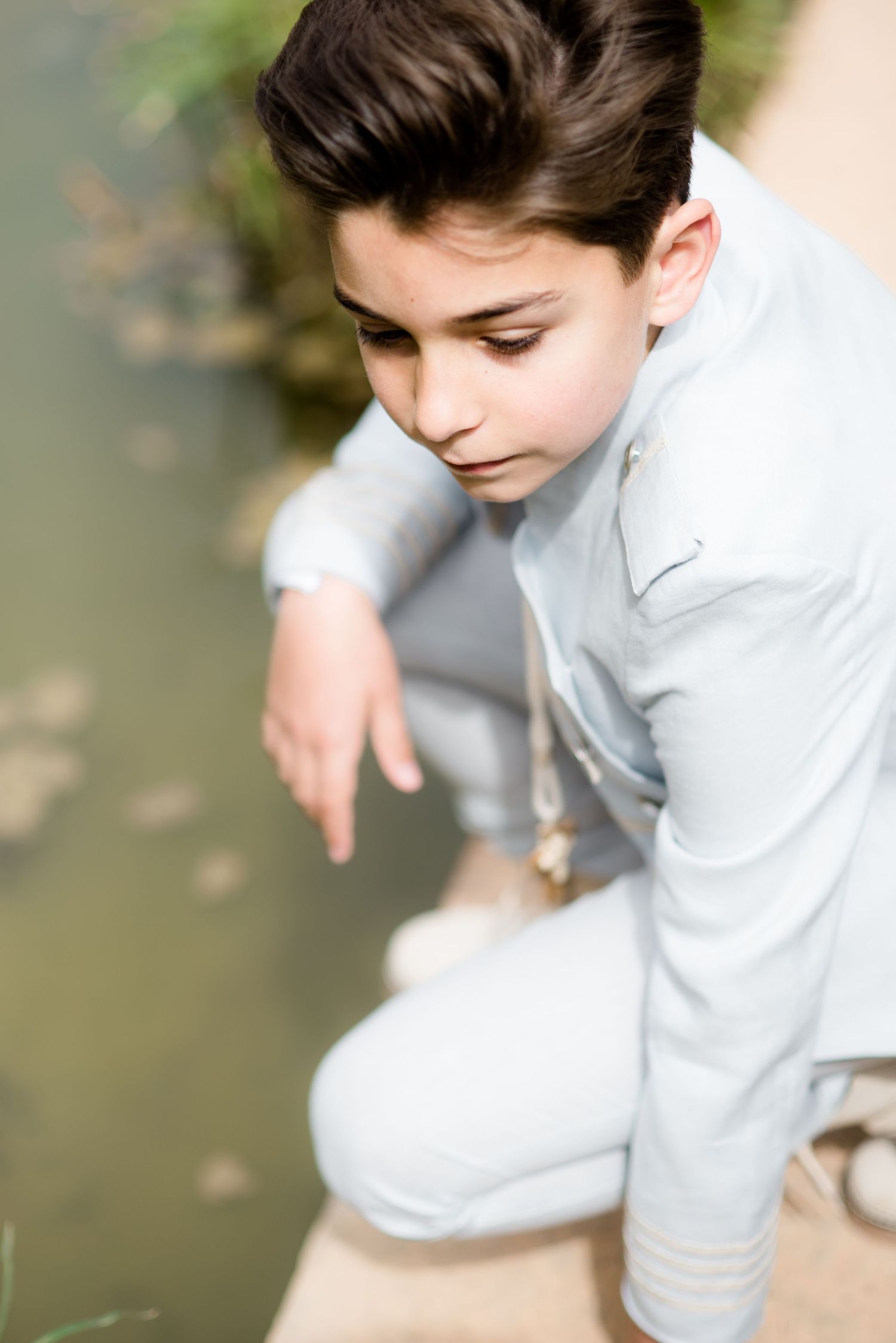 A young boy in a light gray outfit crouches by a pond, pointing at the water.