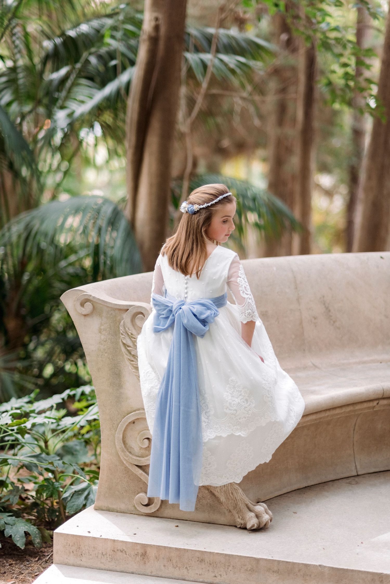 Young girl in white lace communion dress with blue sash bow sits on stone bench in garden.