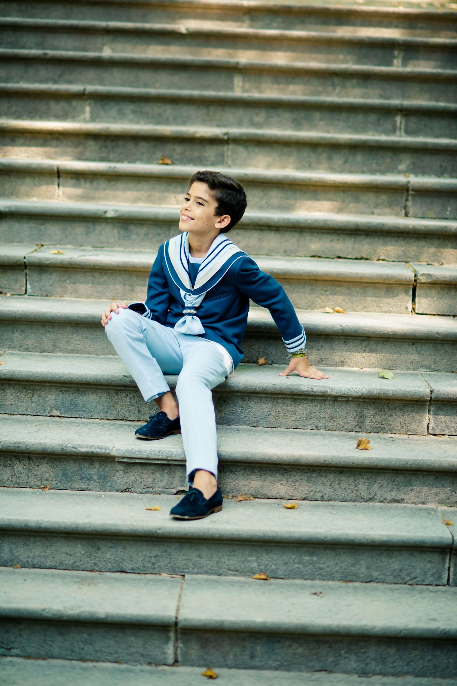 Boy in navy sailor outfit sitting on stone steps outdoors, smiling upward.