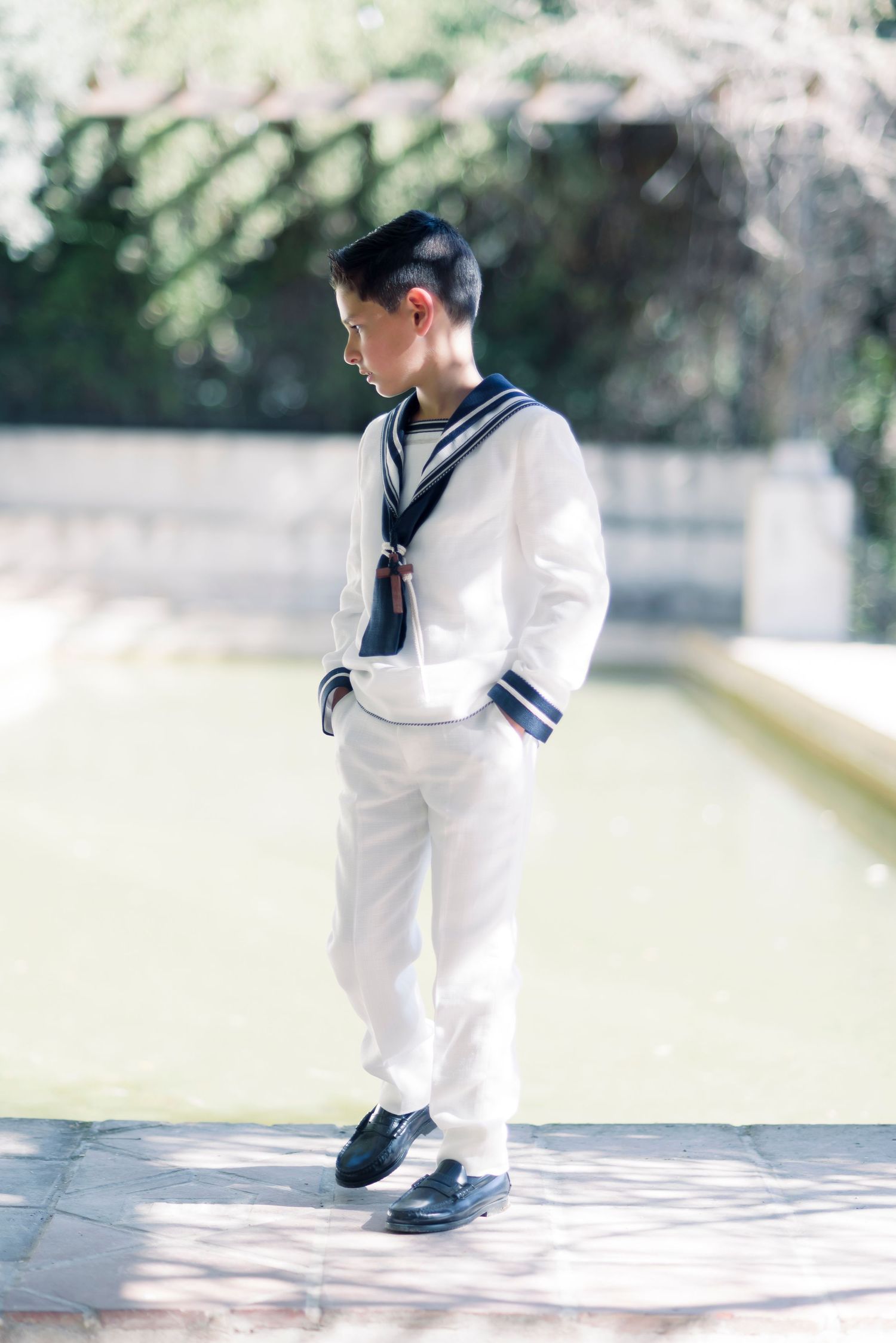 Boy in white sailor communion outfit stands pensively beside garden fountain in sunlight.