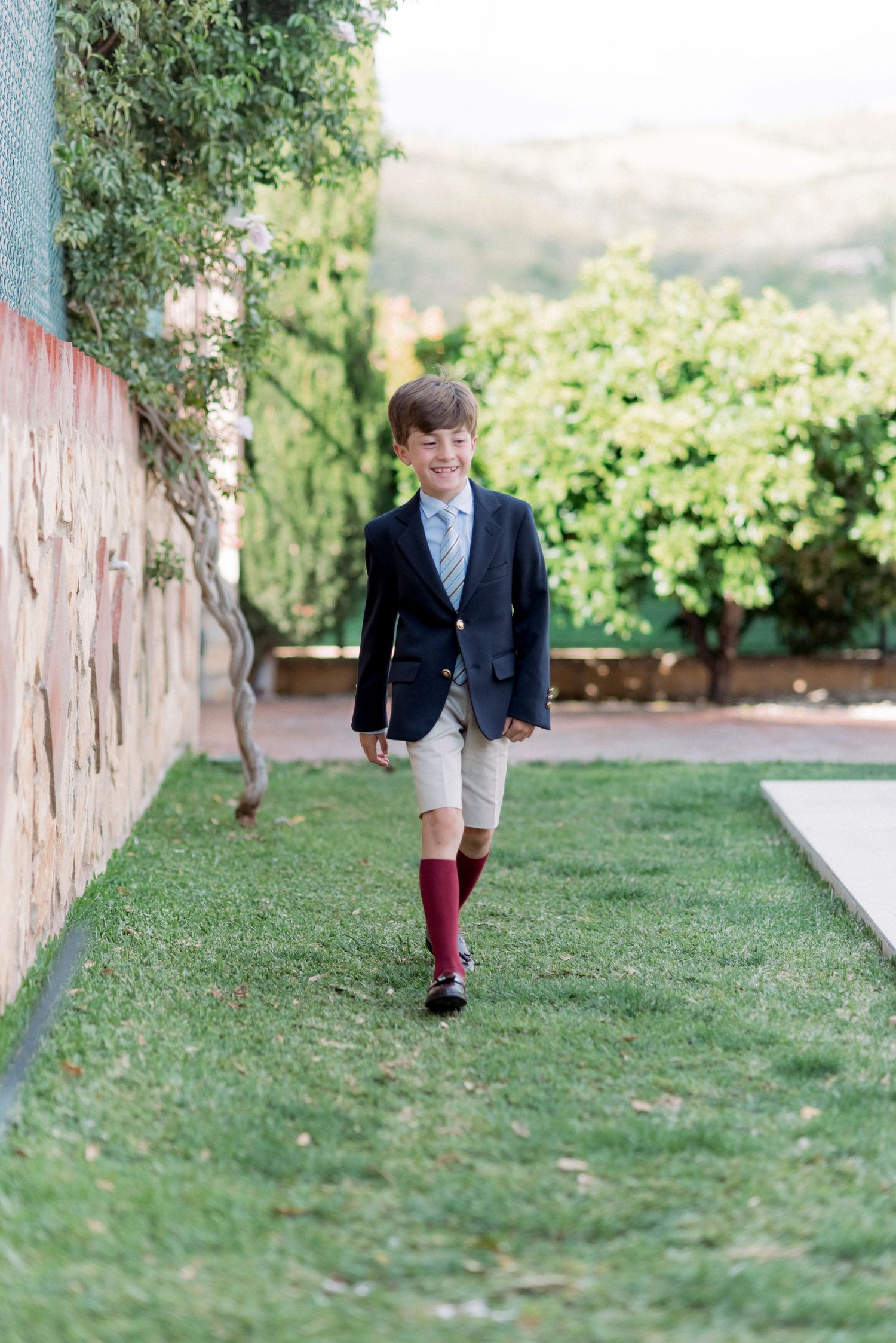 Boy in navy blazer and khaki shorts walking on grass path beside garden wall.