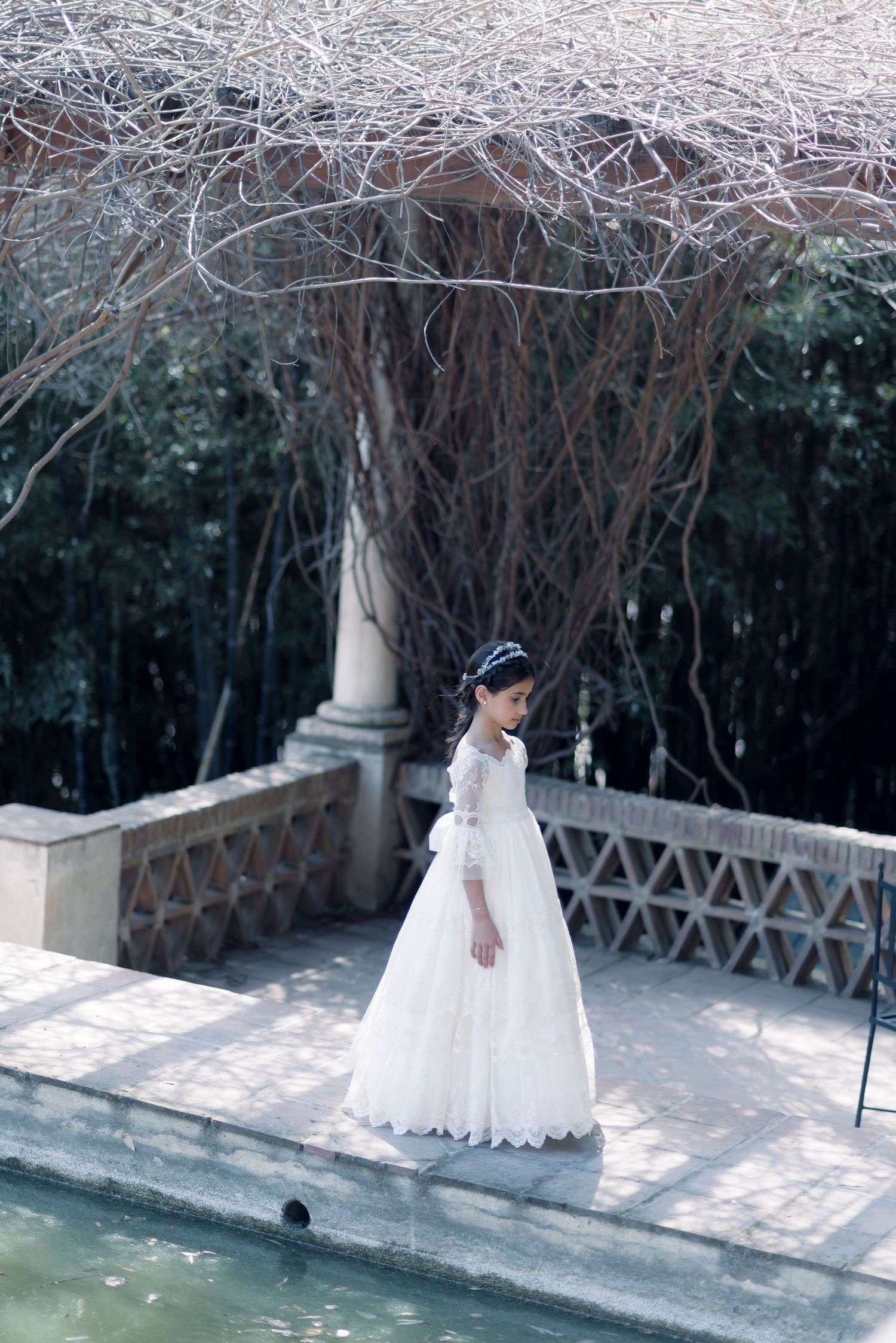 Girl in white lace communion gown walks beside garden pond with bare tree backdrop.