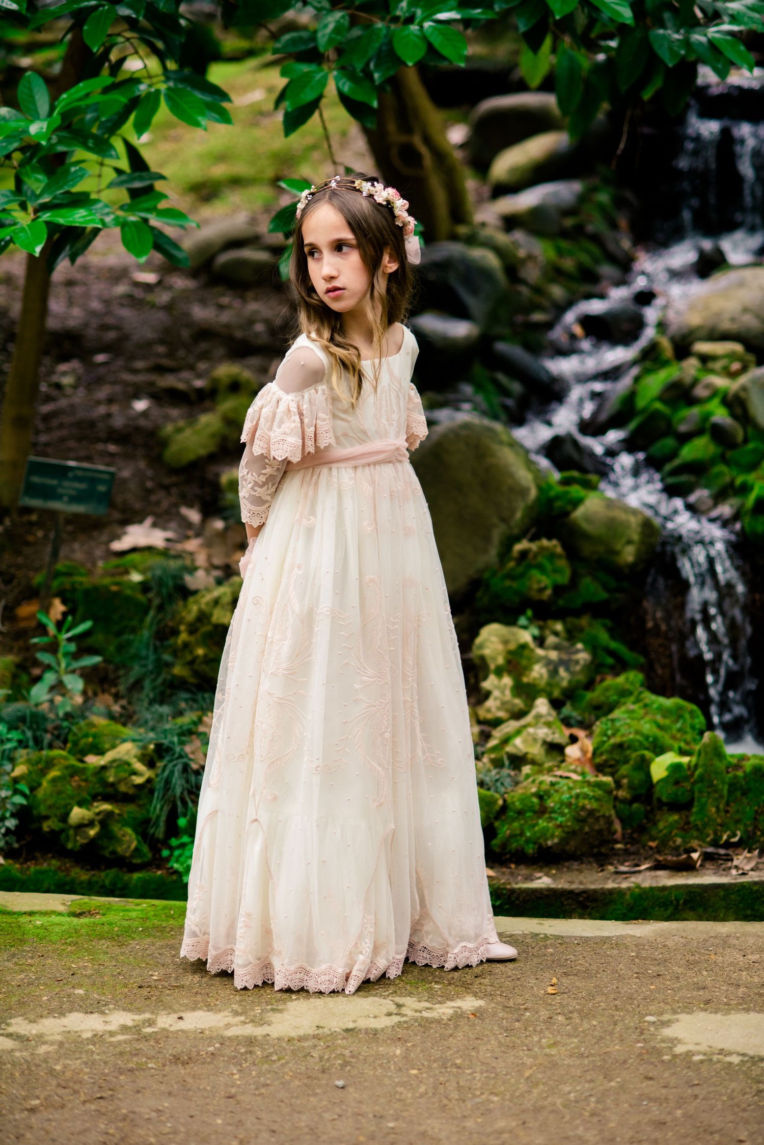 Girl in blush lace communion dress with floral crown by waterfall.