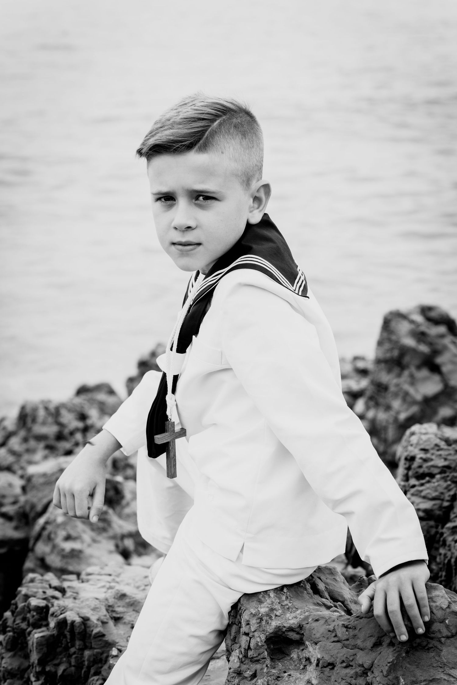 Black and white portrait of boy in white sailor suit posed on rocky coastal shore.